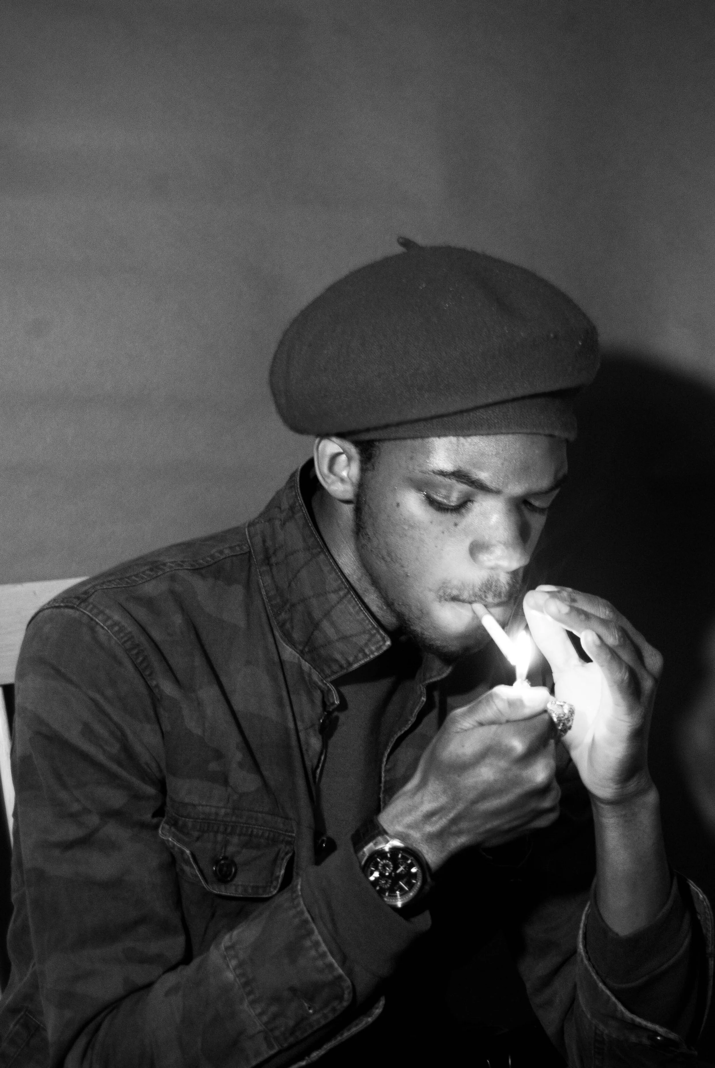 A young man wearing a beret and a jacket is lighting a cigarette with a match in a dimly lit room.