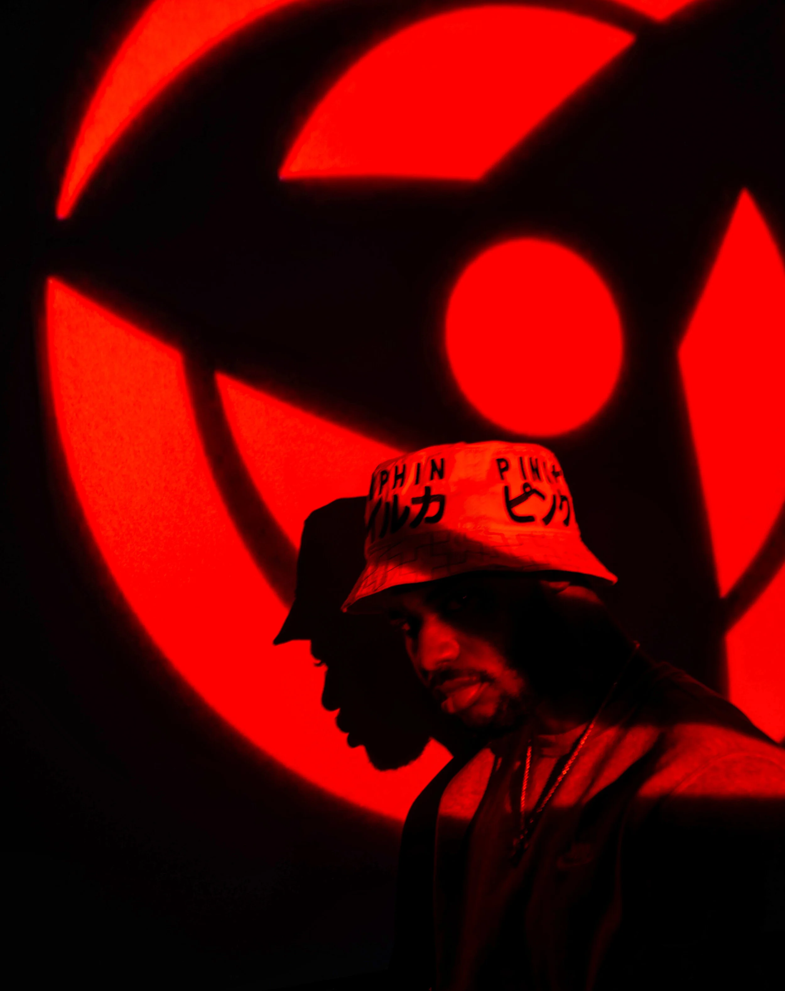 Two people standing in front of a large red and black glowing logo, with the person in the foreground wearing a bucket hat with writing on it and sticking out his tongue.