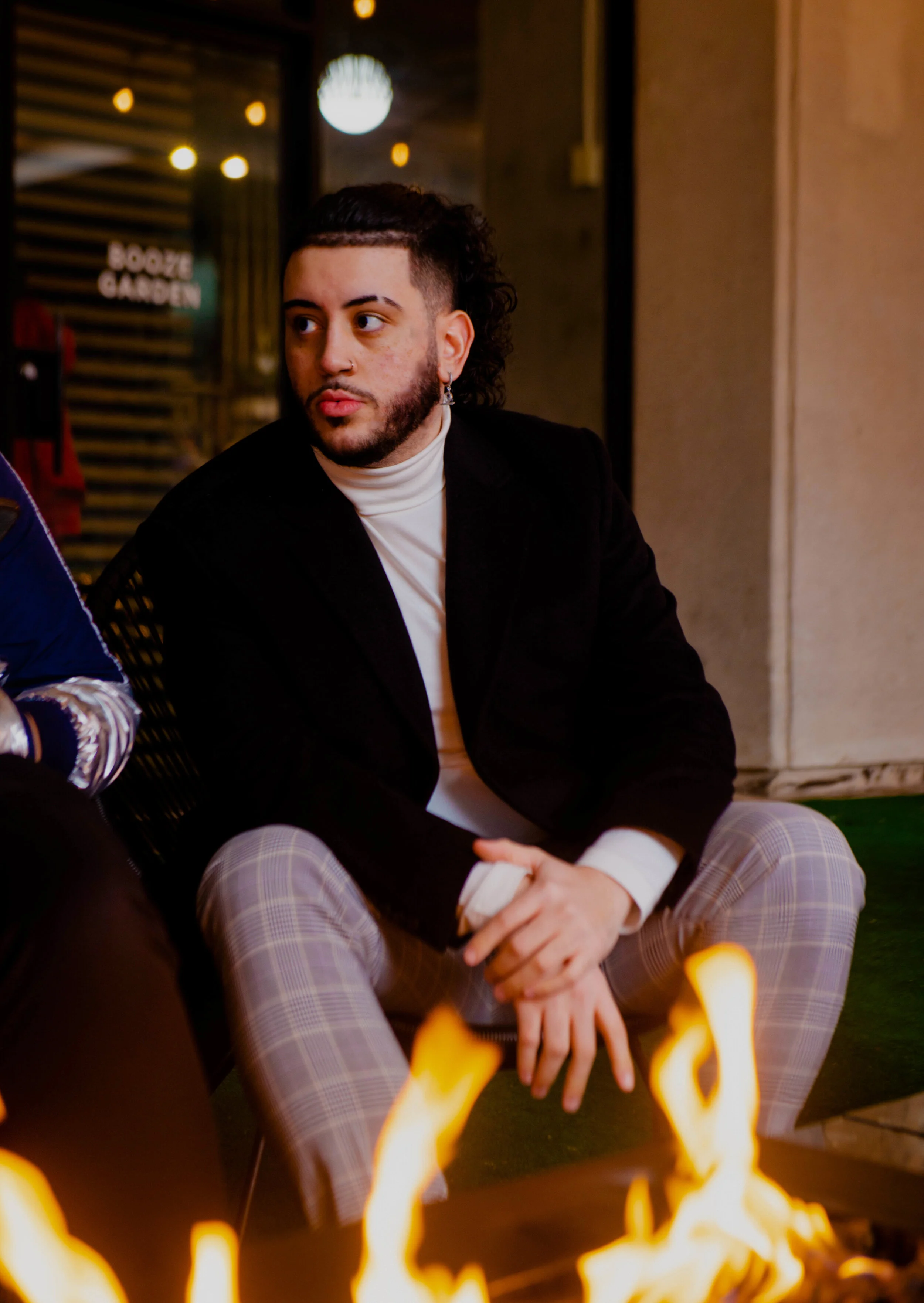 A young man with curly hair, a beard, and earrings sitting by a fire at night, wearing a black blazer, white turtleneck, and plaid pants.