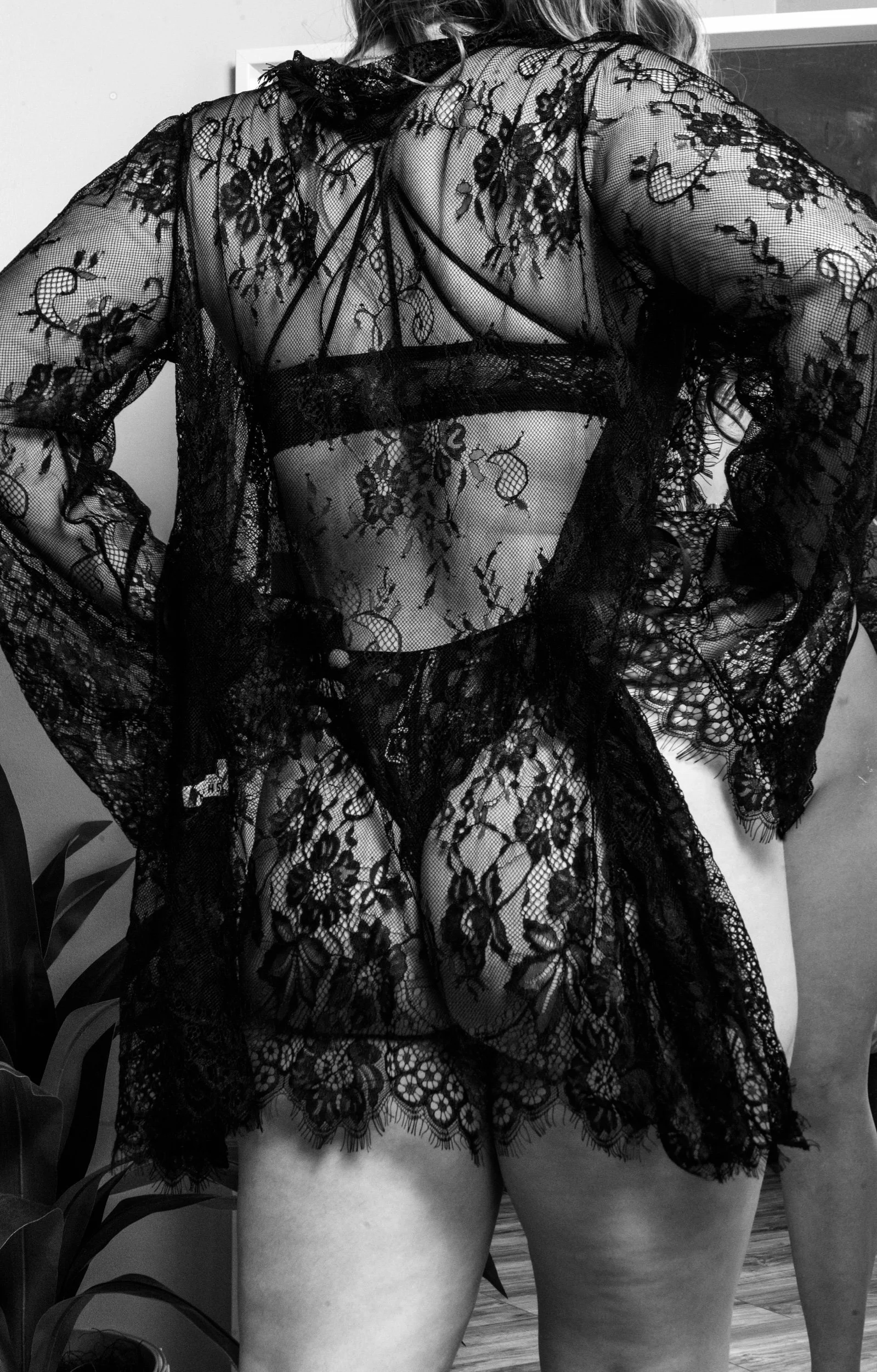 A person wearing black lace lingerie with floral patterns, sitting on a chair with their back to the camera. The lingerie is see-through, revealing the person's skin underneath.