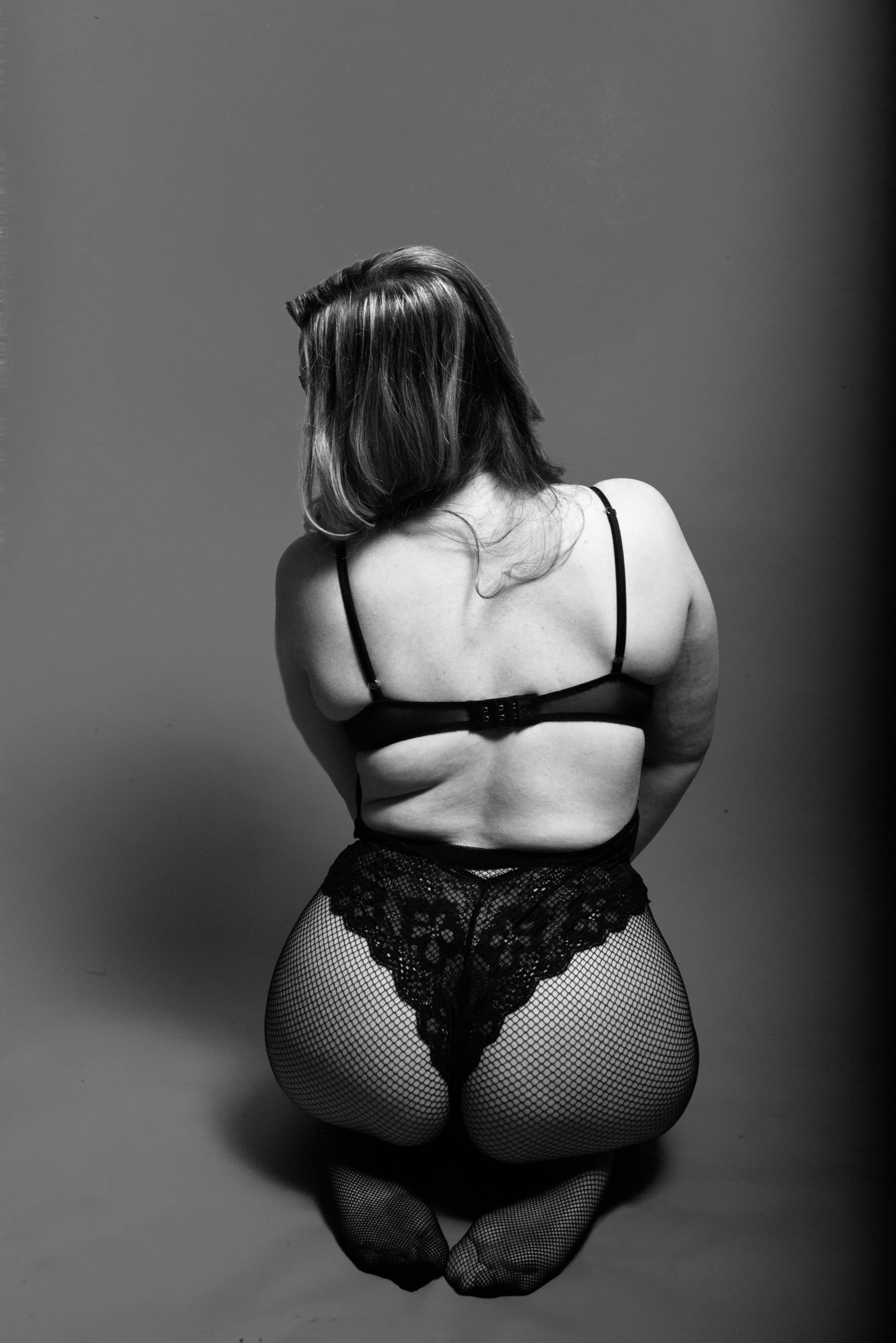 A woman in lingerie, fishnet stockings, and lace underwear kneeling with her back to the camera.