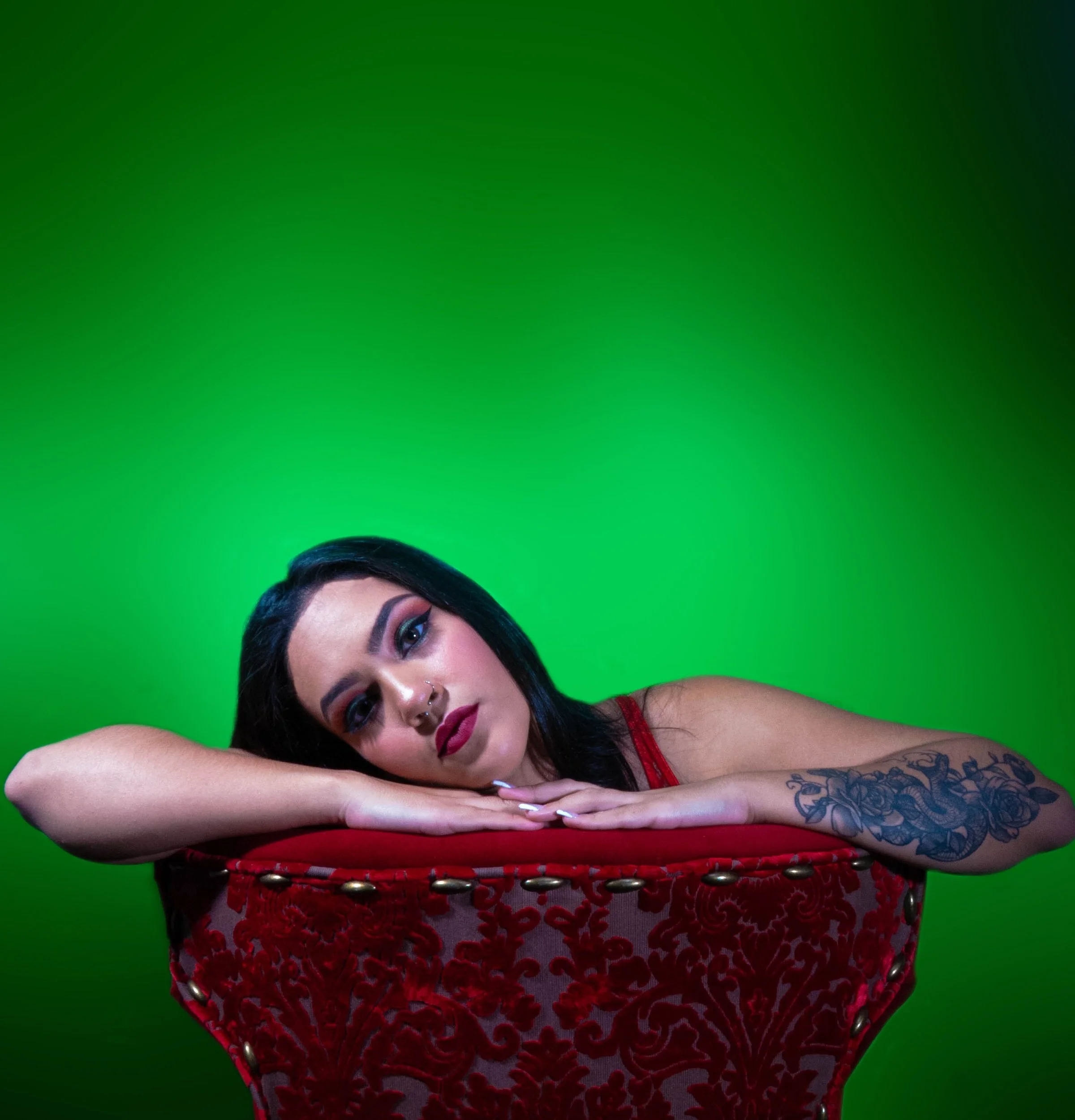 A woman with dark hair and tattoos resting her head on her arms on a red, ornate chair against a green background.