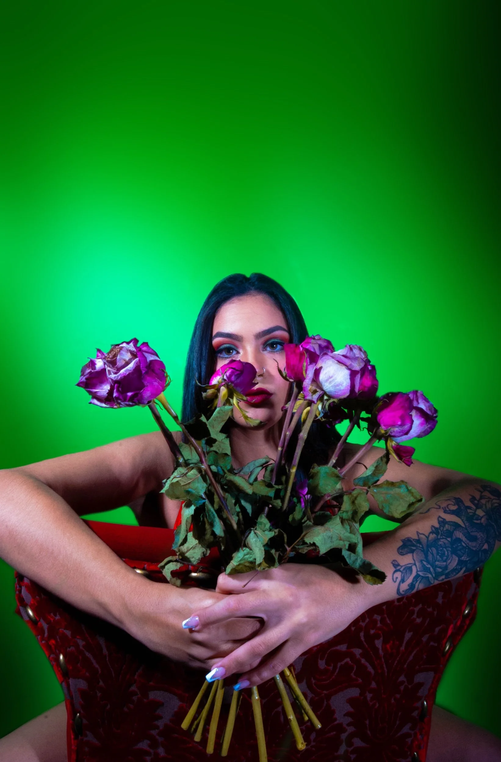 A woman with dark hair, colorful eye makeup, and tattoos on her arms holding a bouquet of wilted purple roses in front of a green background.