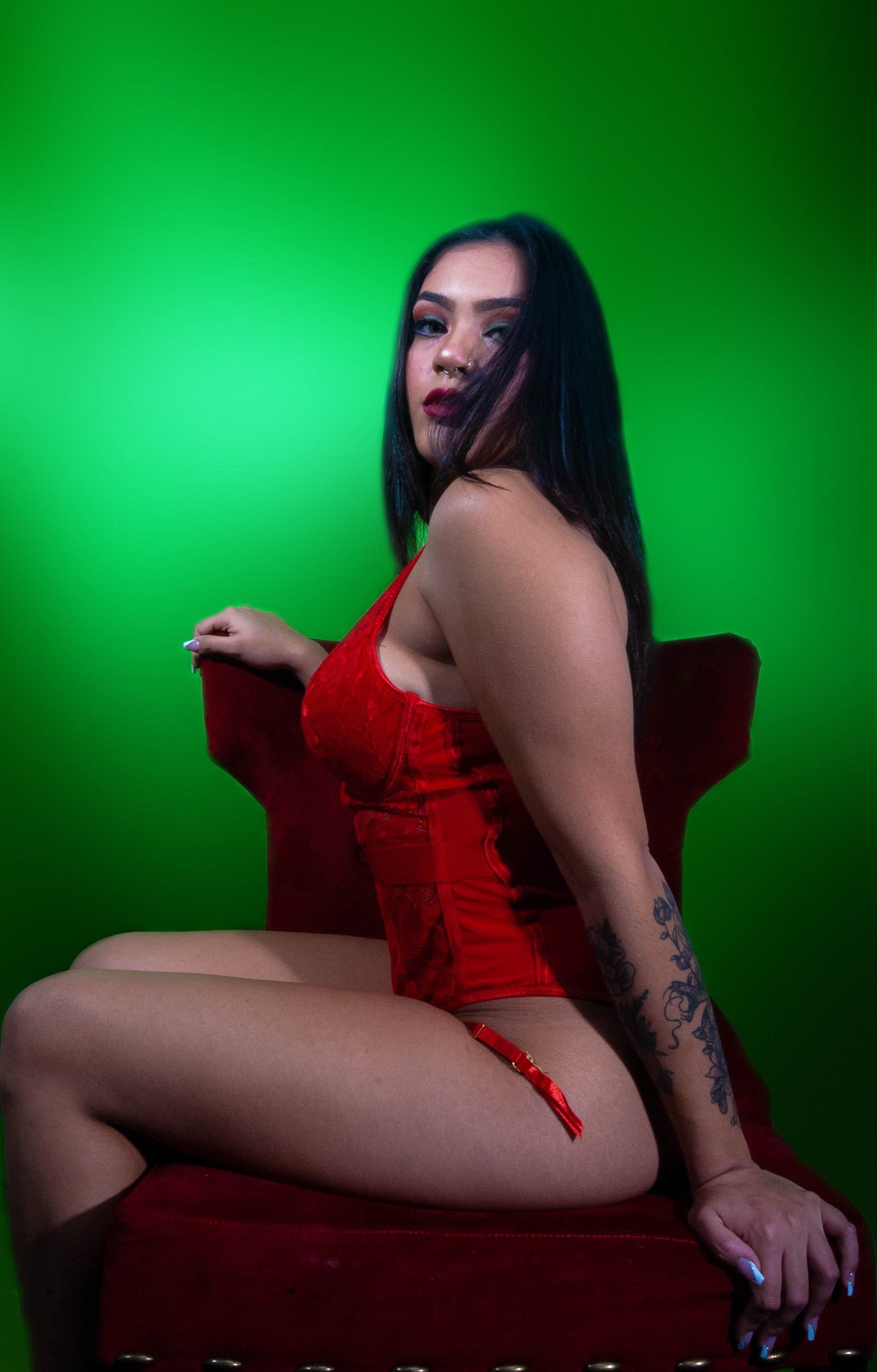 A woman with dark hair, wearing a red corset, sitting on a red chair with a green background.
