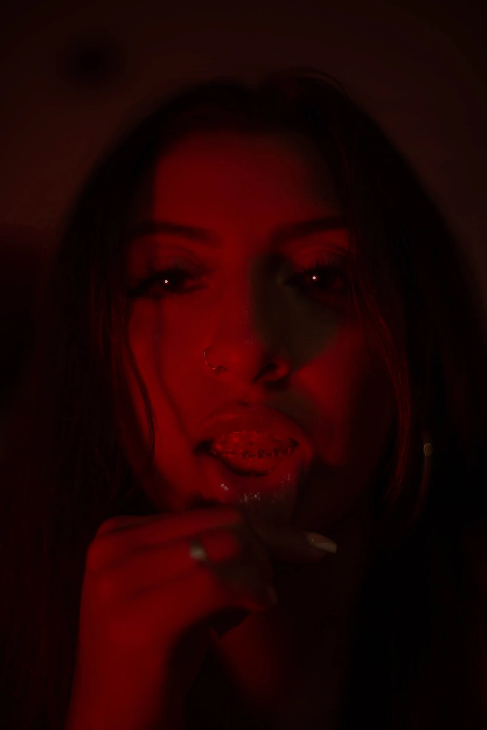 A woman with dark hair and makeup, holding her chin with her hand, illuminated by red light.