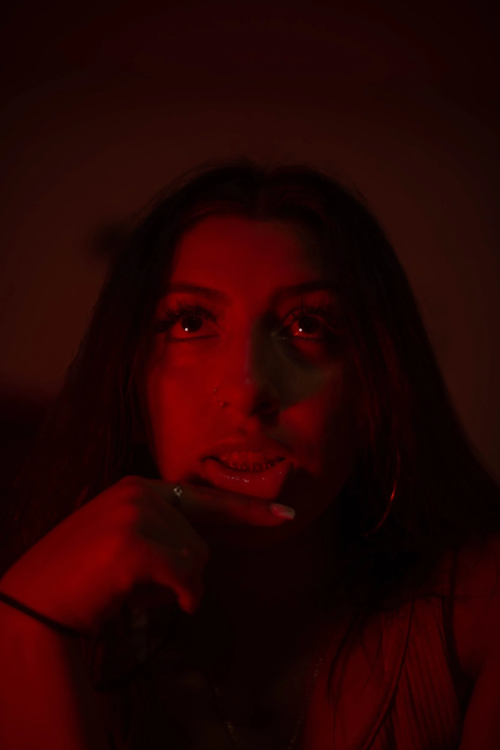 A woman with dark hair, wearing hoop earrings and a nose ring, under red lighting, with her mouth slightly open and hand near her chin.