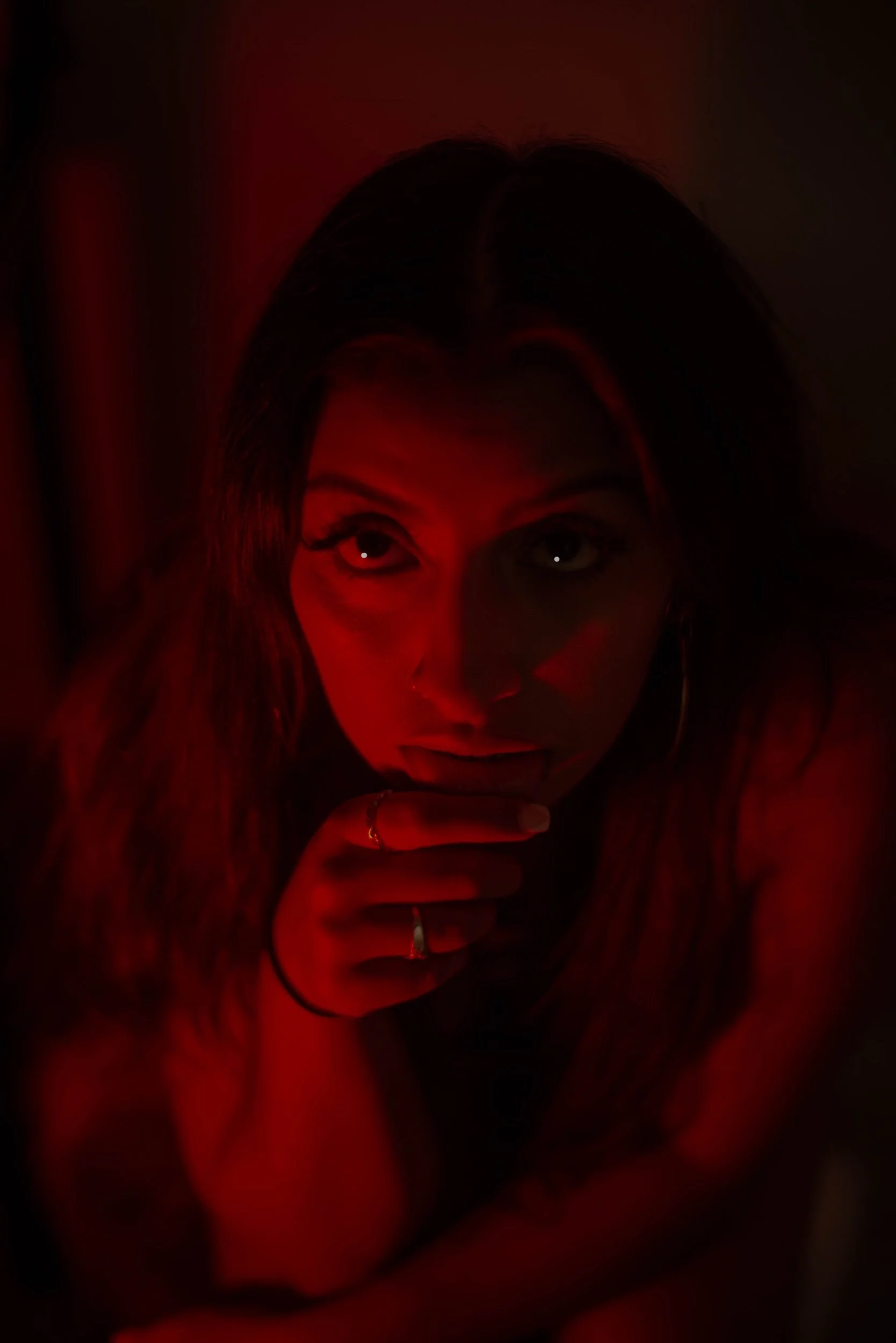 A woman with long hair and hoop earrings is looking up at the camera with an intense expression, illuminated by red lighting.