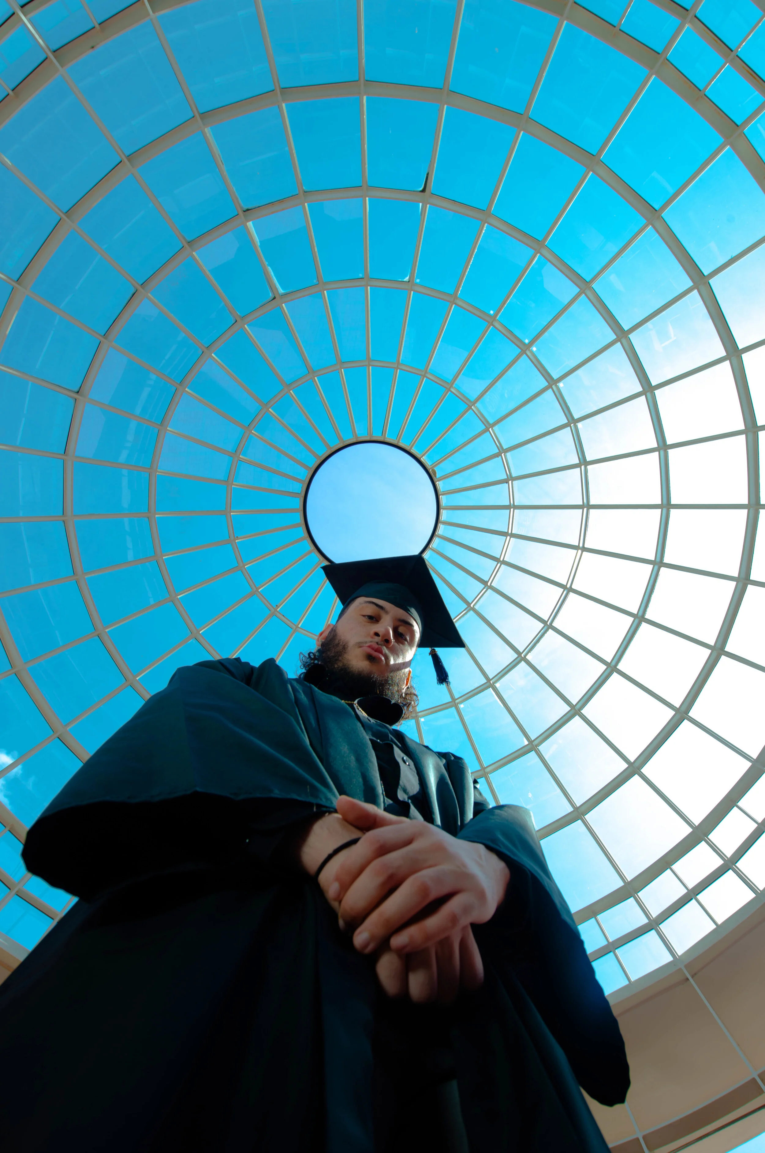 Low-angle photo of a man in a graduation cap and gown standing under a large glass dome ceiling.