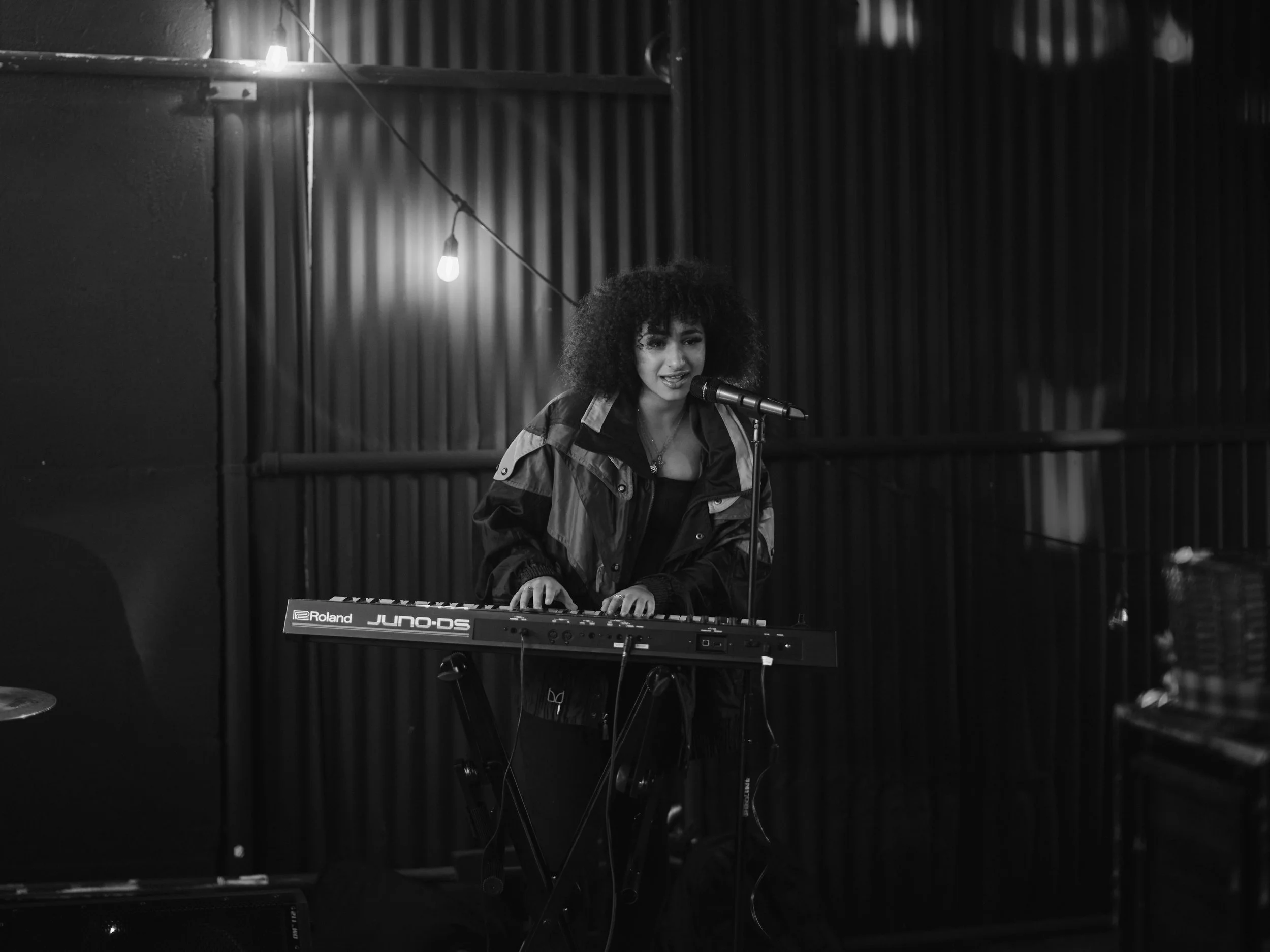 A woman with curly hair performing with a keyboard and microphone in a dimly lit space.