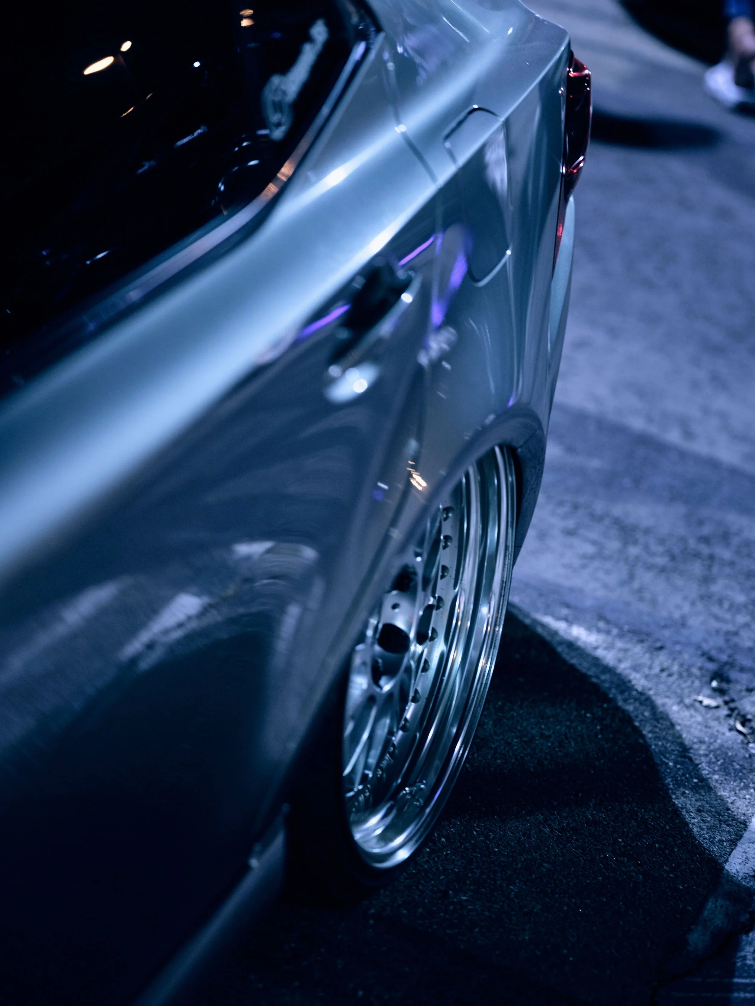 Close-up of a lowered silver sports car with deep-dish wheels, parked on asphalt at night with blue and purple lighting.