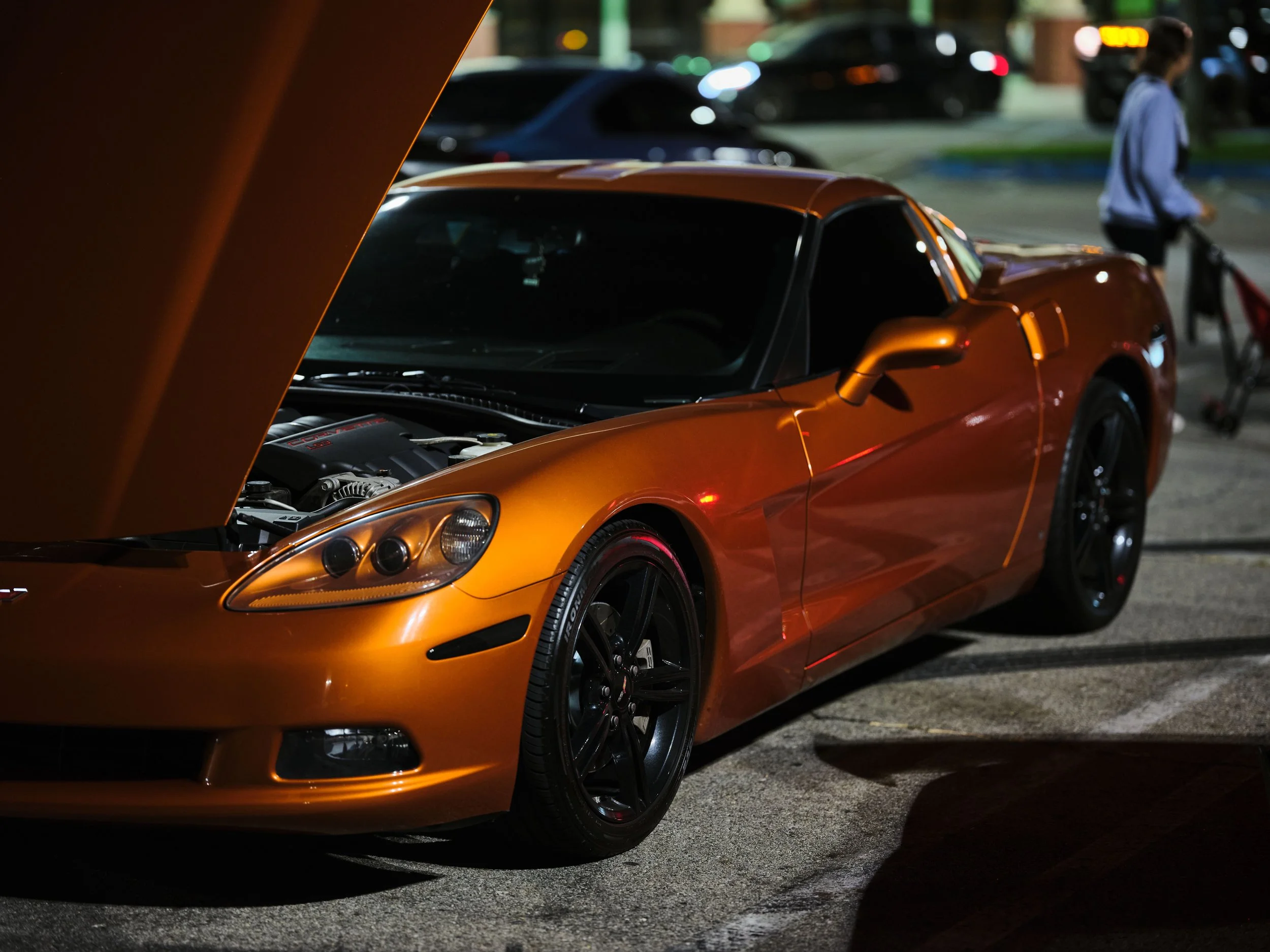 Orange sports car with open hood parked at night in a parking lot, with a person and shopping cart in the background.