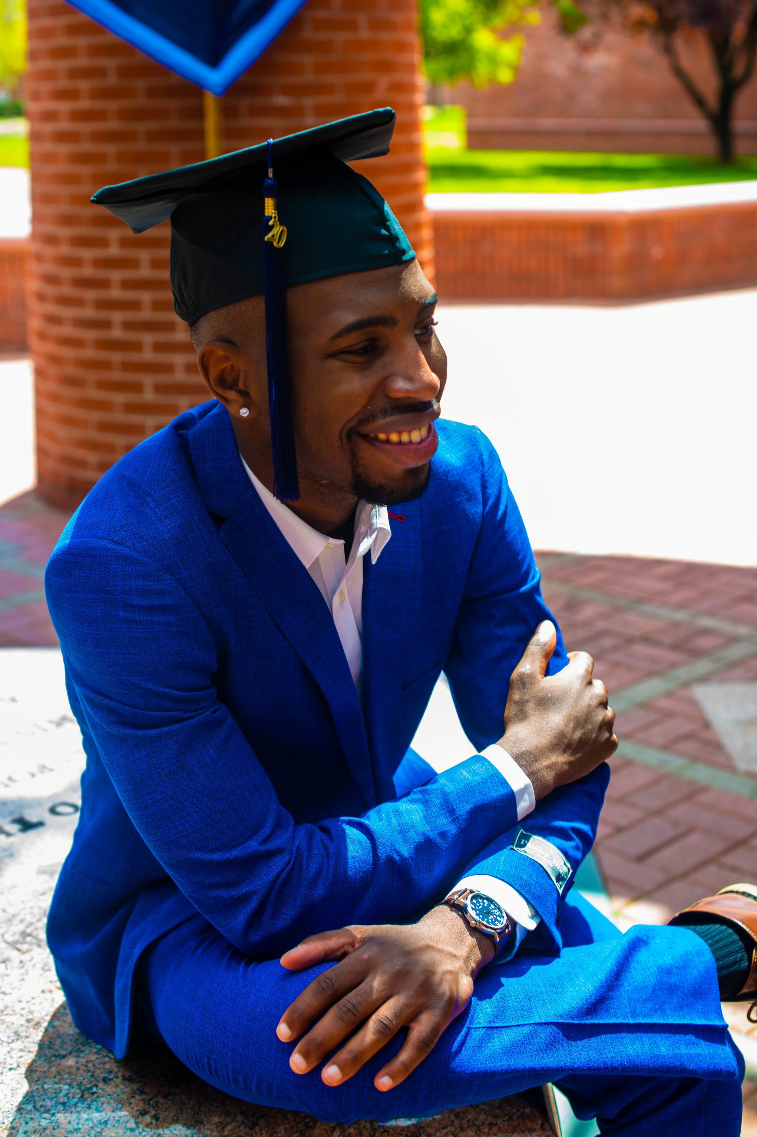 A young man in a bright blue suit and graduation cap sitting outdoors and smiling, with a brick structure and trees in the background.