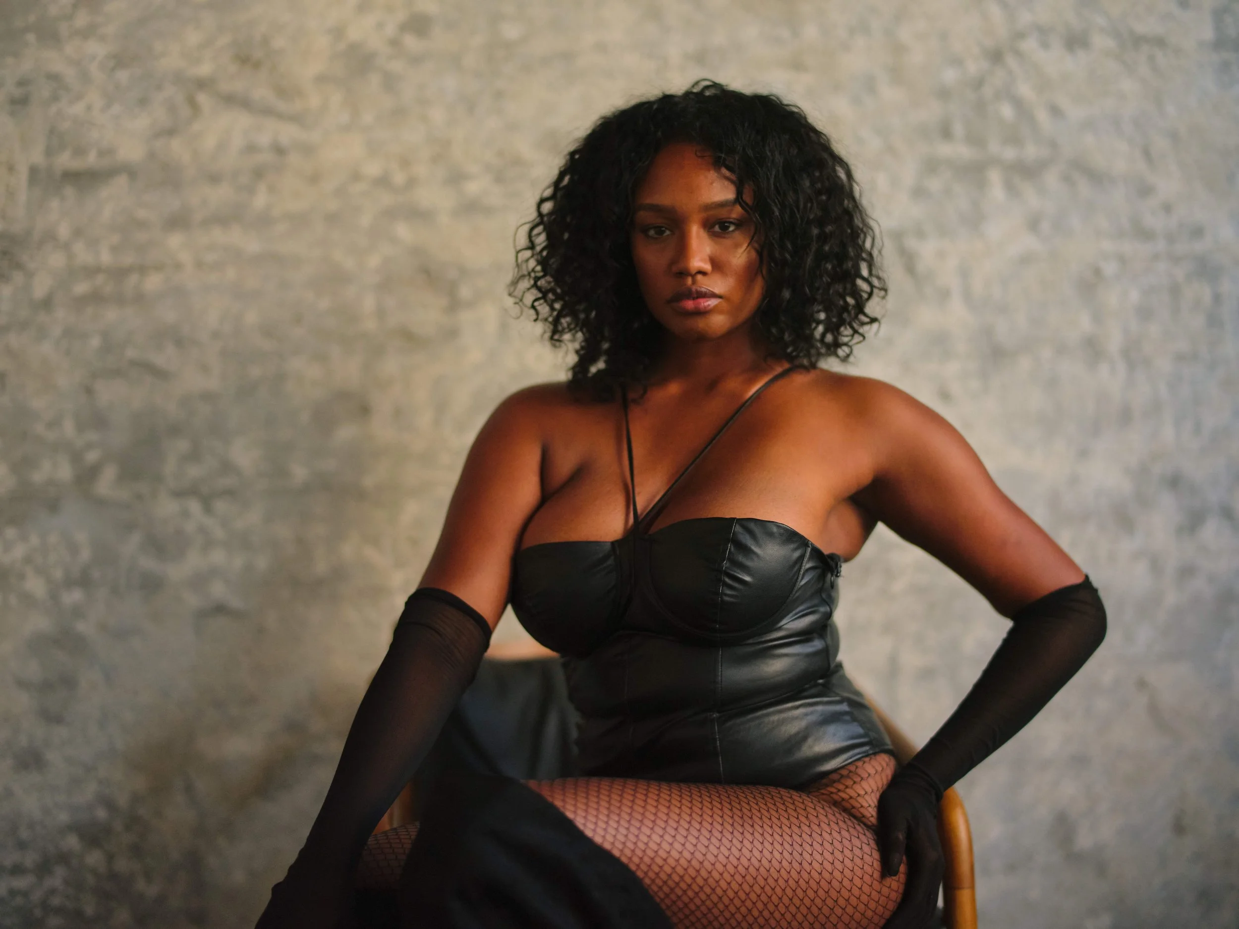 A woman with curly black hair wearing a black leather dress, fishnet stockings, and sheer black gloves seated on a wooden chair against a textured gray wall.