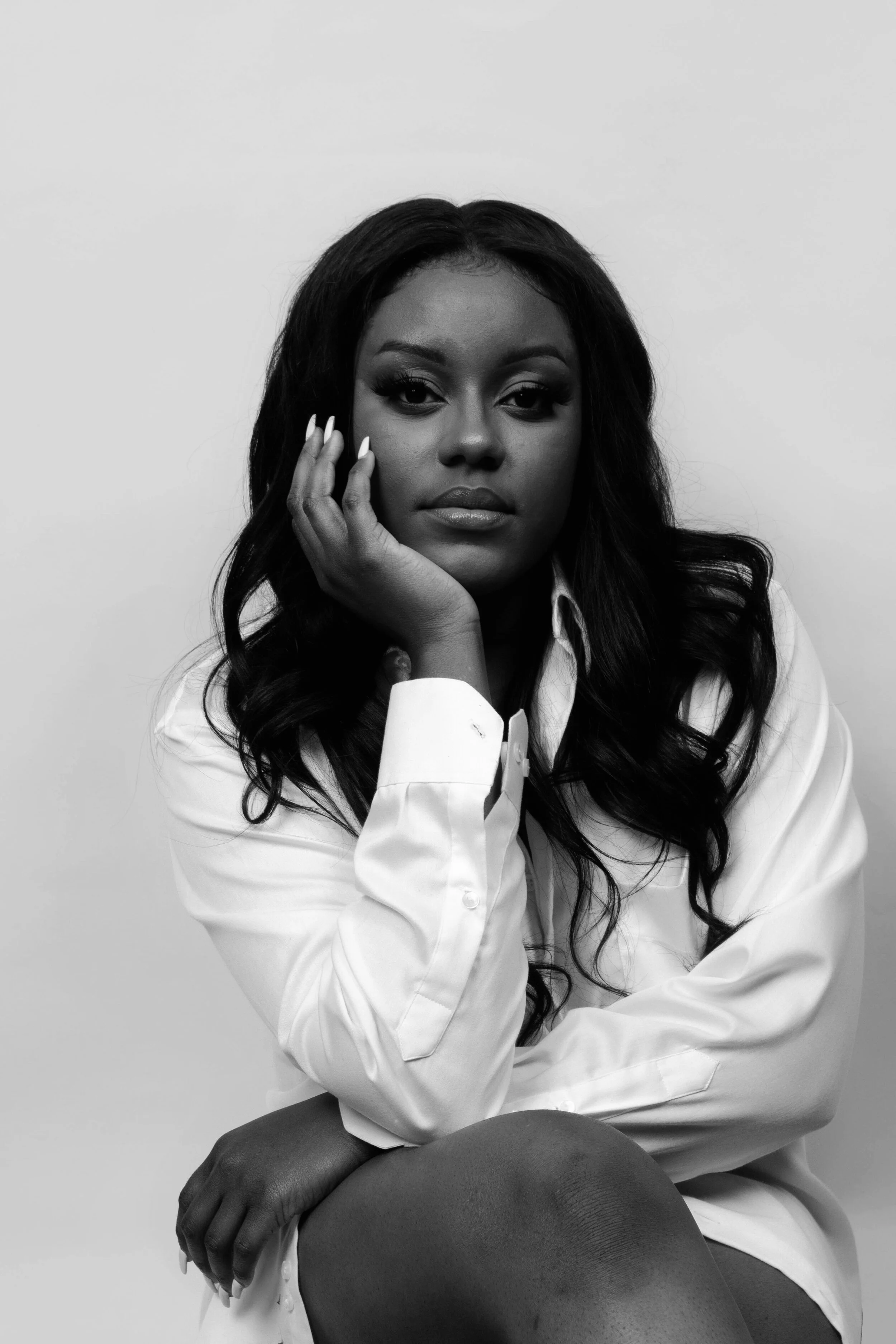 Black and white portrait of a woman with long, wavy hair, wearing a white shirt, sitting with her hand resting on her face, against a plain background.