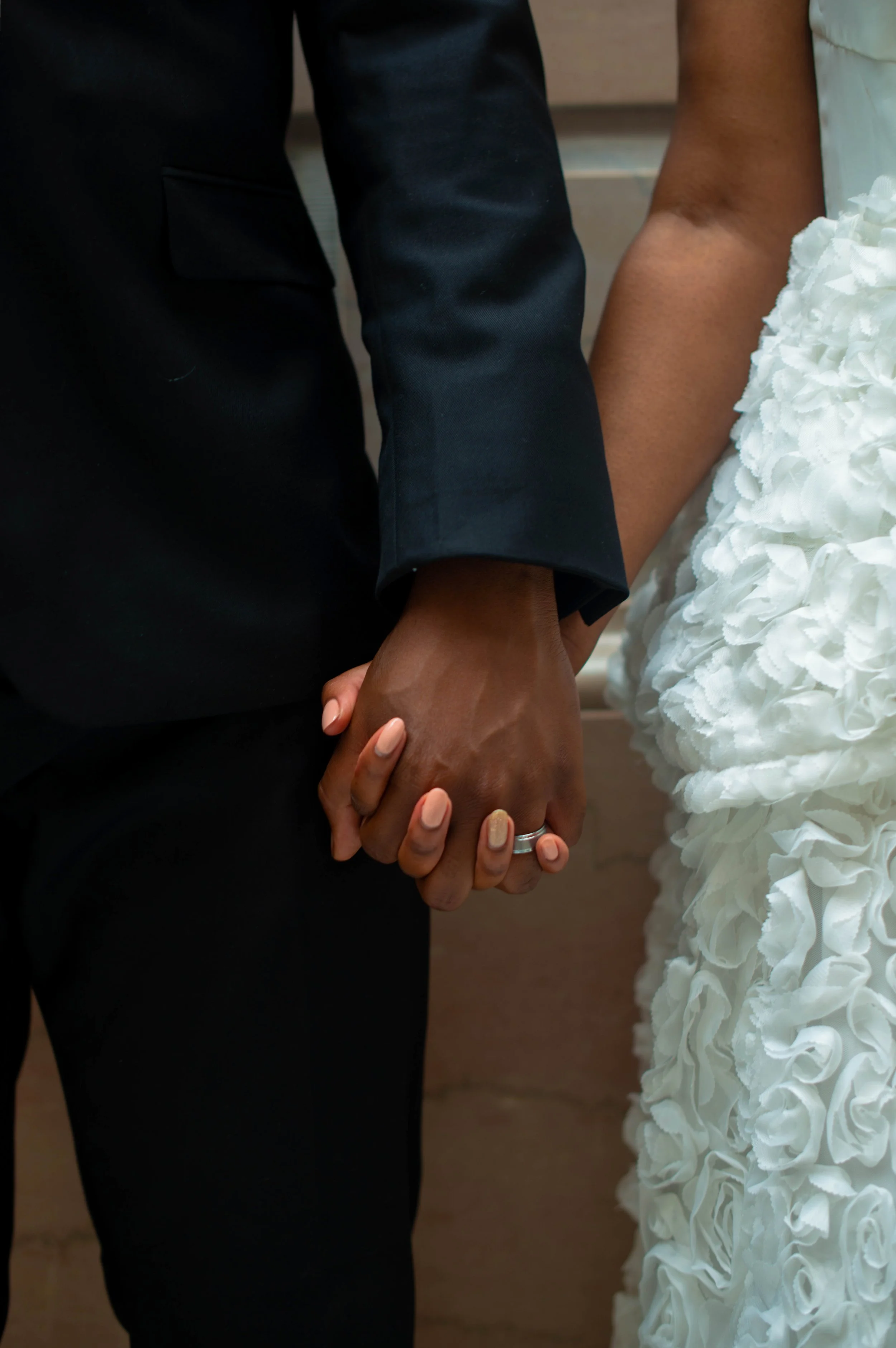 Close-up of a couple holding hands during a wedding, with the bride wearing a white dress with textured floral details and the groom in a black suit, showing wedding rings on their fingers.