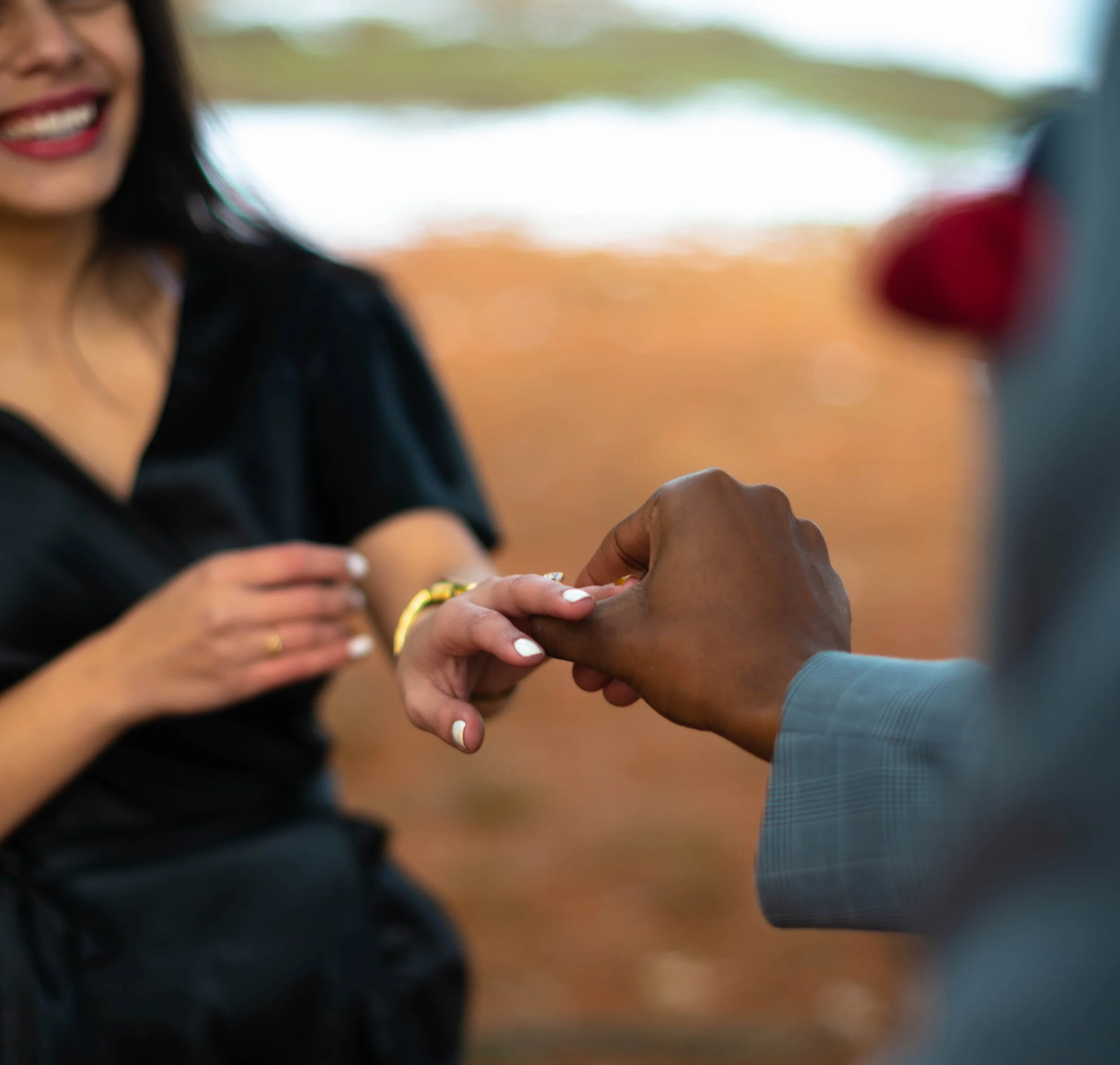 A woman smiling as a man gently holds her hand, possibly during an outdoor event or celebration, with a blurred background of orange and green hues.