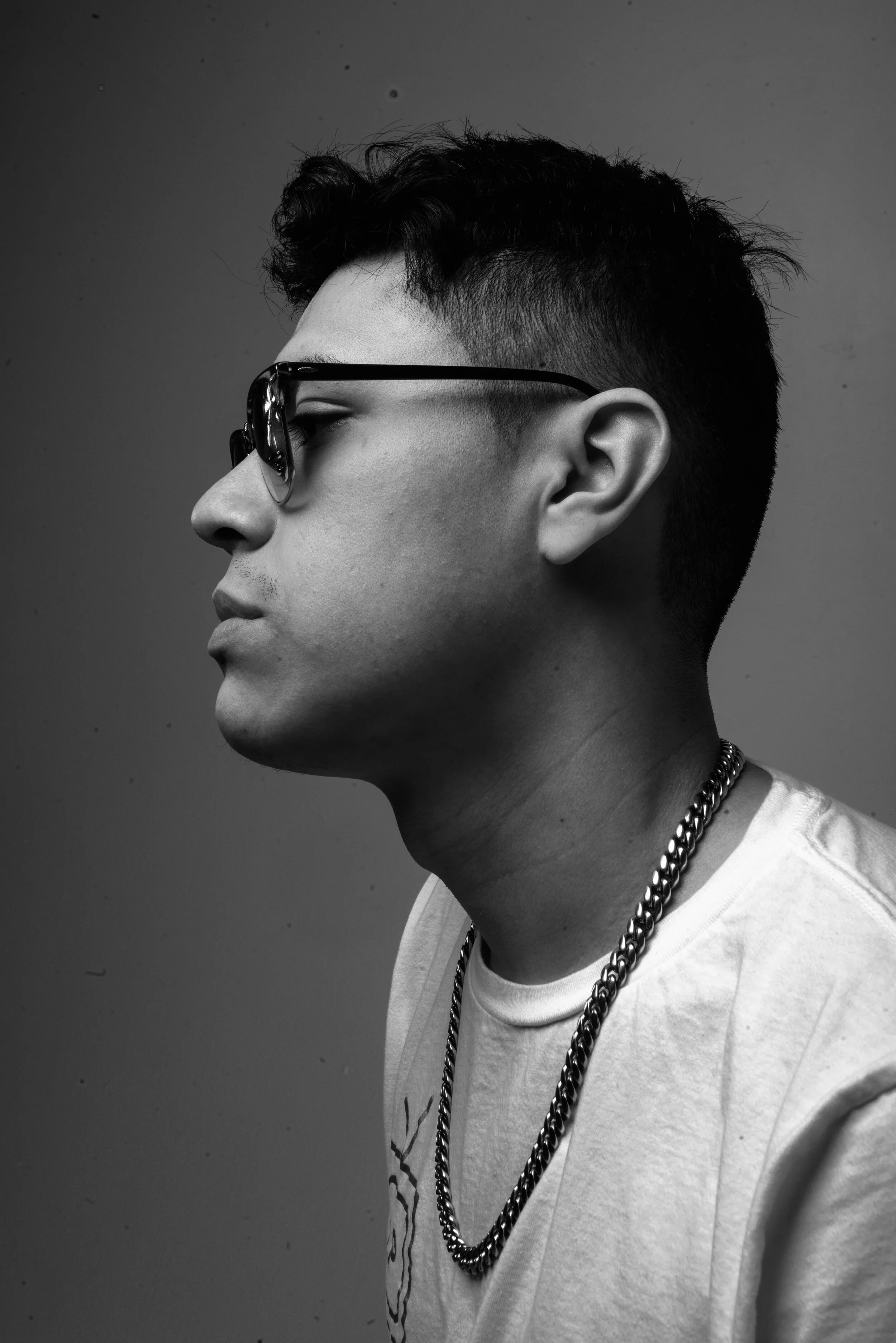 Black and white side profile of a young man with styled hair, wearing sunglasses, a chain necklace, and a T-shirt, against a plain background.