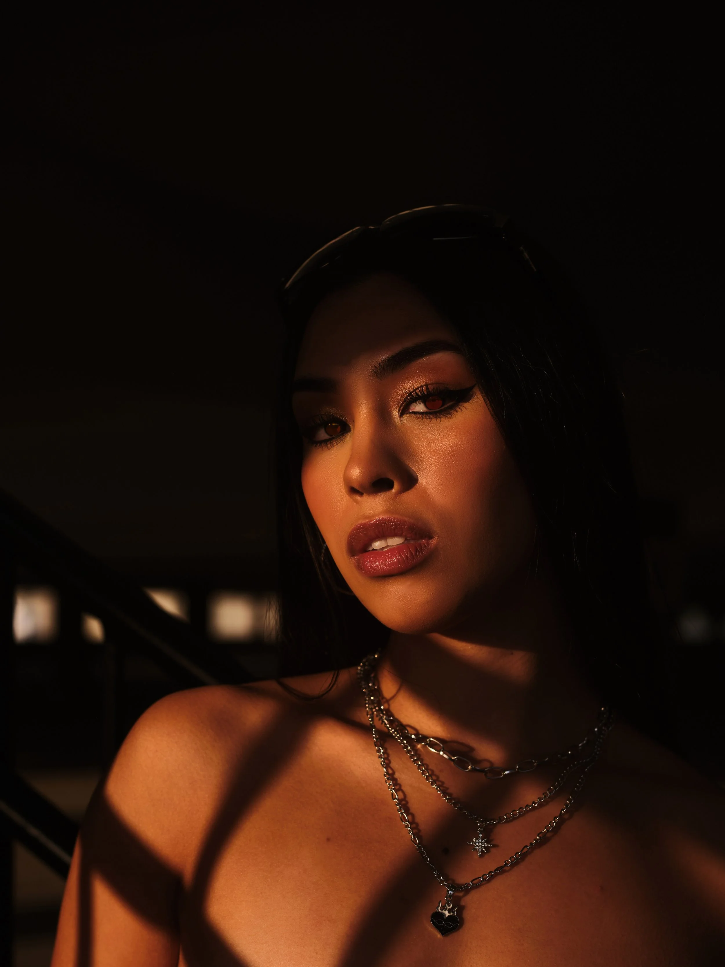 A woman with dark hair and makeup, wearing layered silver necklaces, is partially in shadow with warm lighting highlighting her face and shoulders.