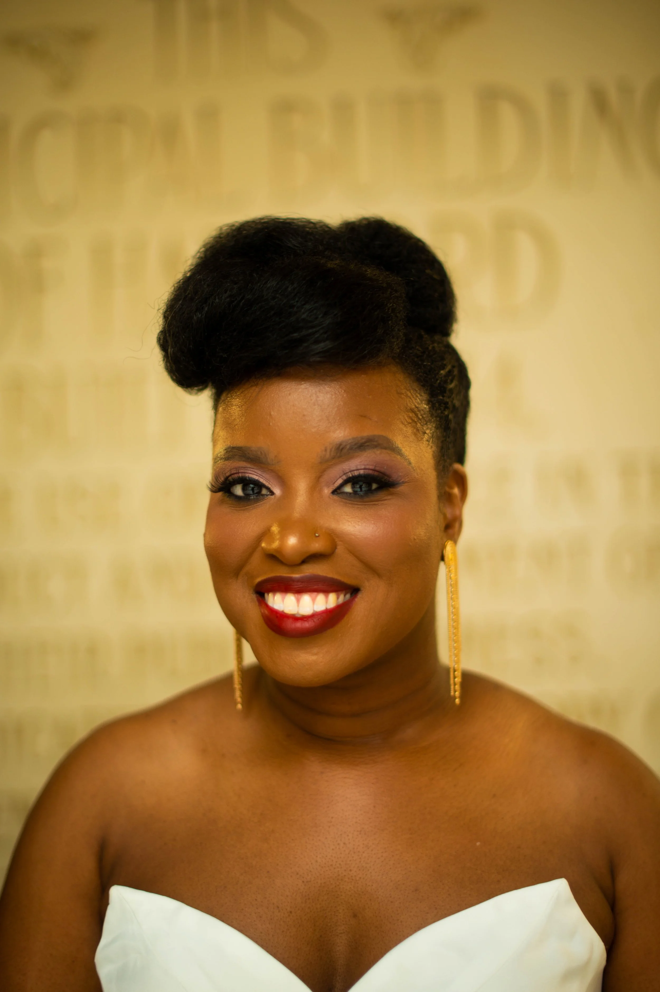 A woman with dark skin, styled hair, and red lipstick smiling at the camera, wearing long earrings and a strapless white dress.