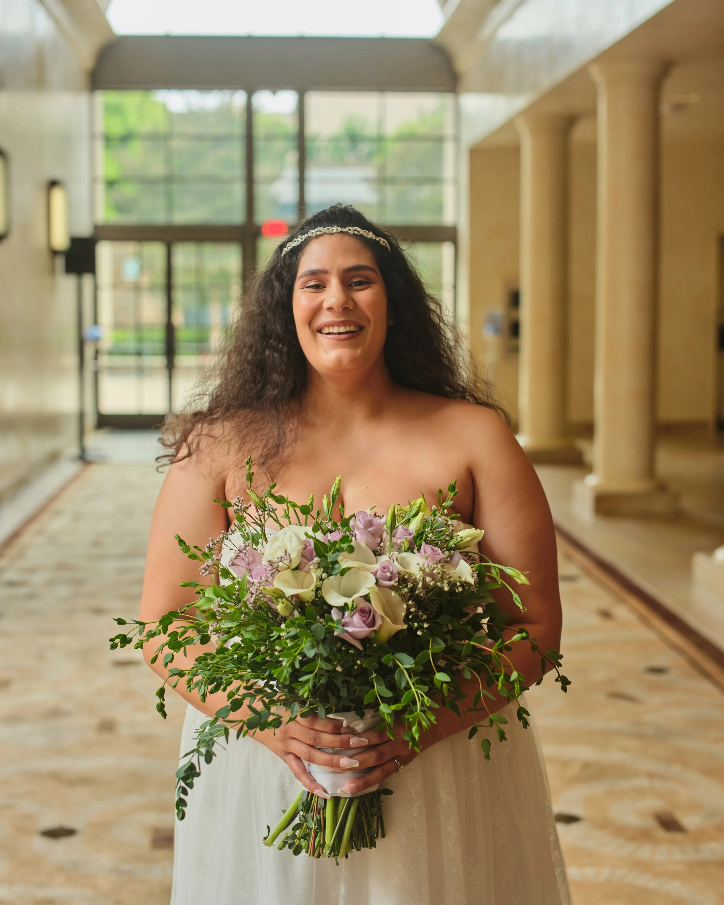 A smiling bride holding a bouquet of white and purple flowers inside a building with large windows and beige walls.