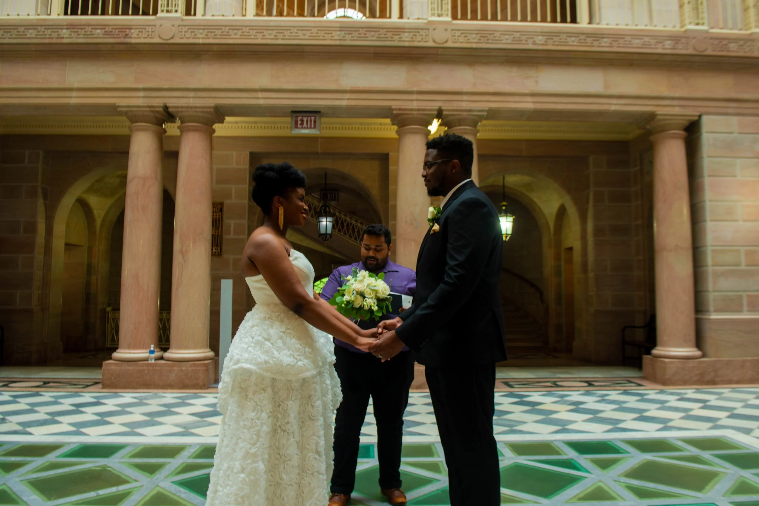 A couple gets married indoors, holding hands and facing each other with a officiant in the background. The bride wears a white strapless gown, and the groom wears a black suit with glasses. The officiant is wearing a purple shirt and holding a bouquet of white and yellow flowers.