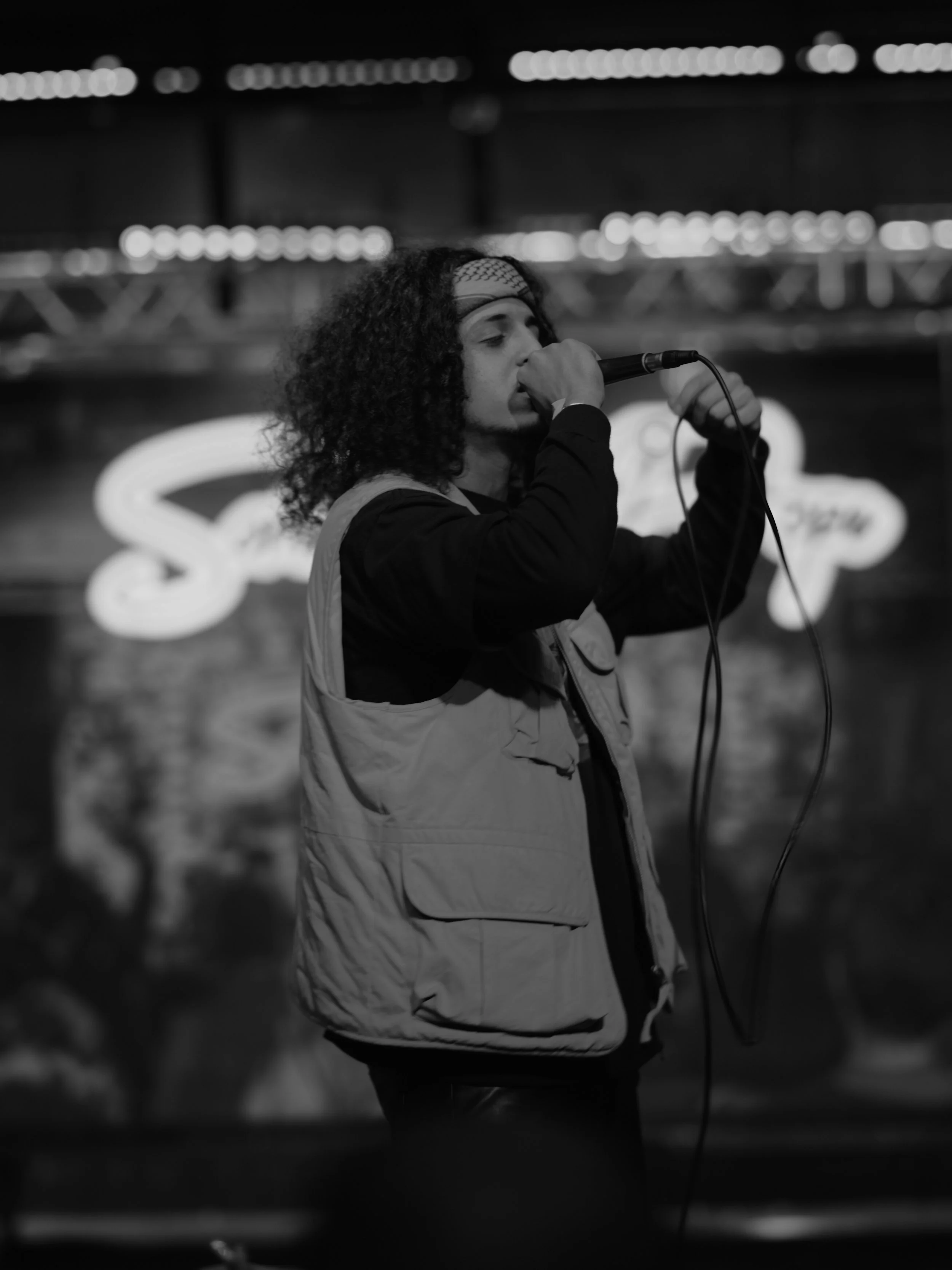A person singing into a microphone on stage in a nightclub or bar with a blurred sign in the background.