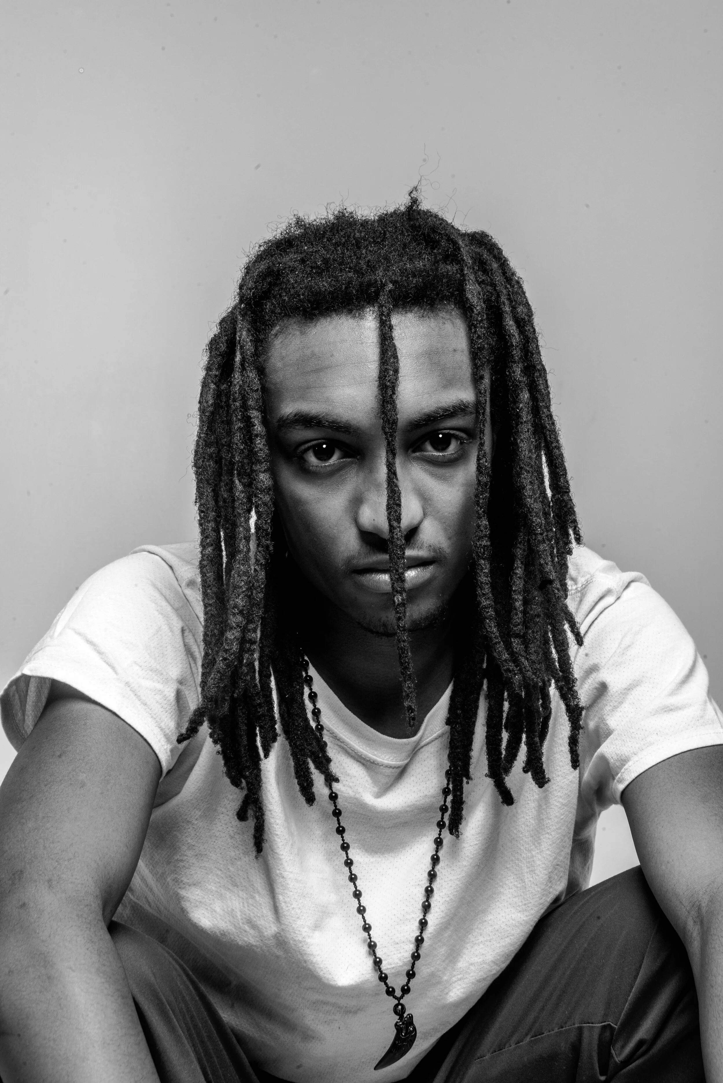 Black and white portrait of a young man with dreadlocks, wearing a white t-shirt and a necklace with a pendant, sitting against a plain background.