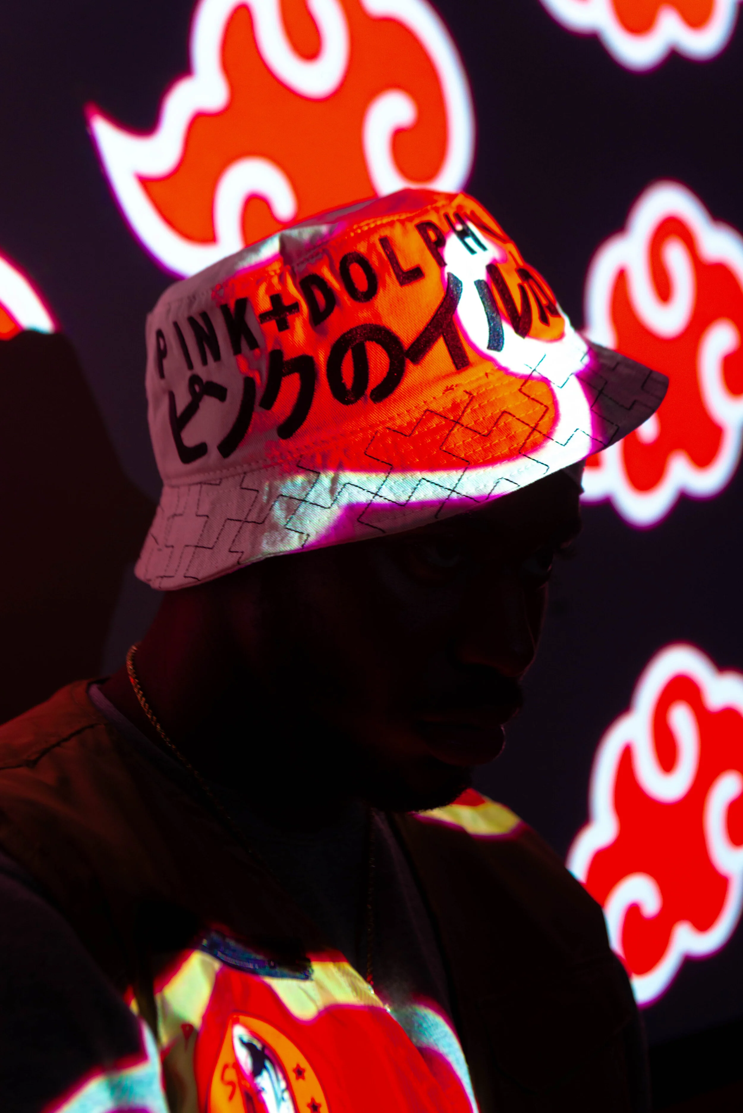 Person wearing a bucket hat with Japanese text and a cartoon graphic, illuminated by colorful projected lights, with red and white cloud designs in the background.