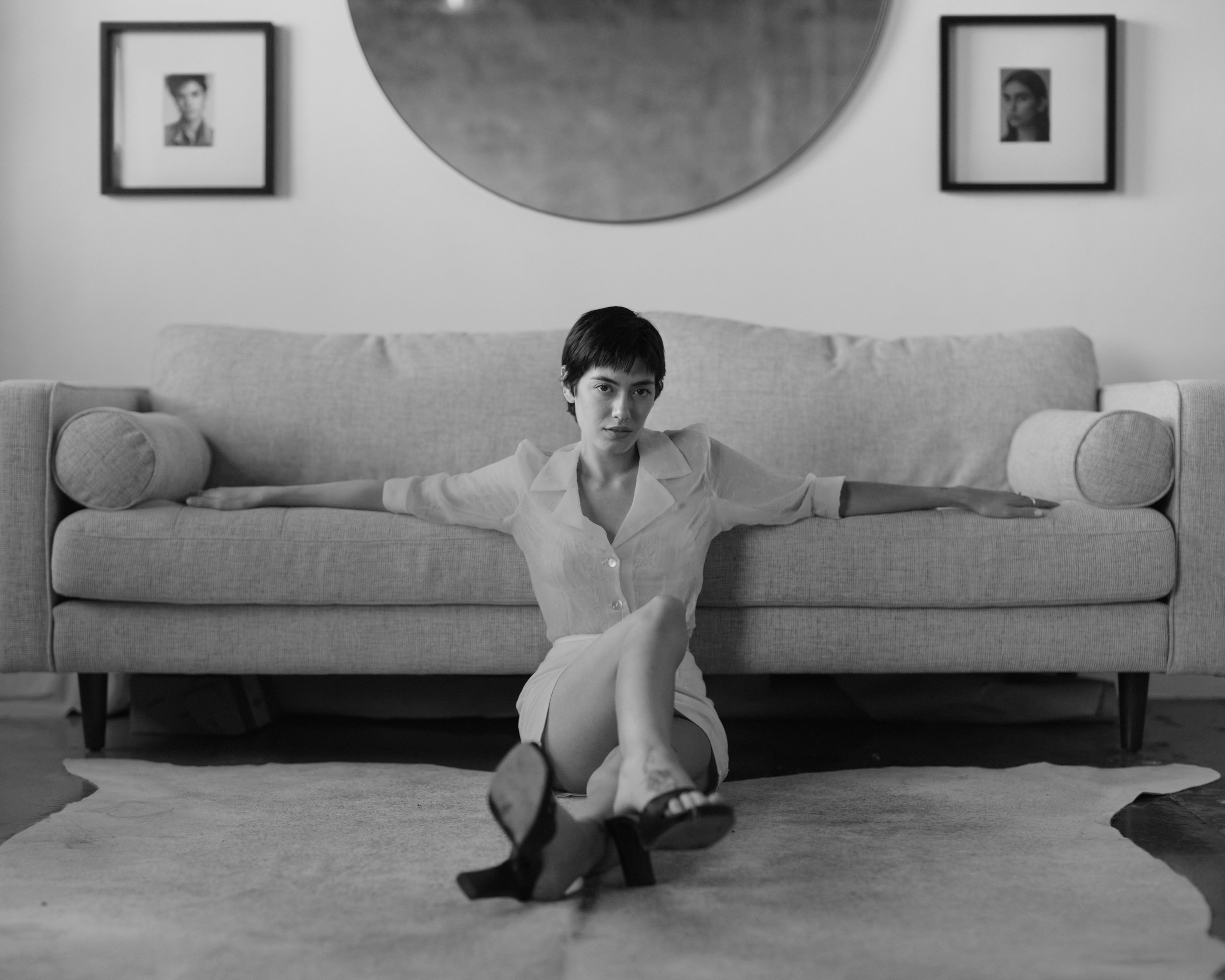 A black and white photo of a woman with short hair sitting on the floor in front of a sofa, wearing a short-sleeved shirt and high heels, with her legs crossed and arms extended along the sofa's back. There are framed photographs hanging on the wall 