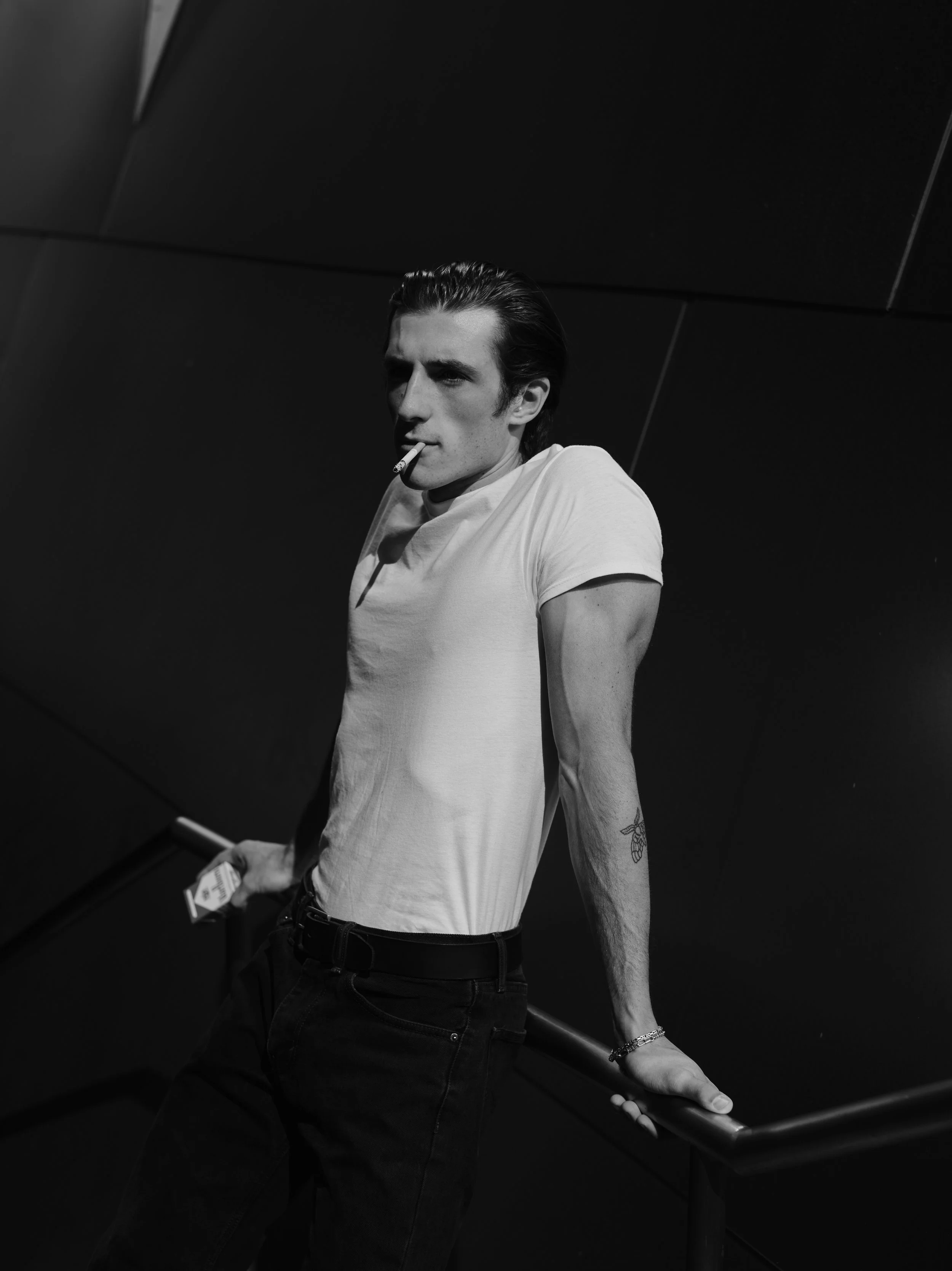 A young man with slicked-back hair wearing a white t-shirt and dark jeans, holding a cigarette in his mouth, standing against a dark background, leaning on a railing with one hand.