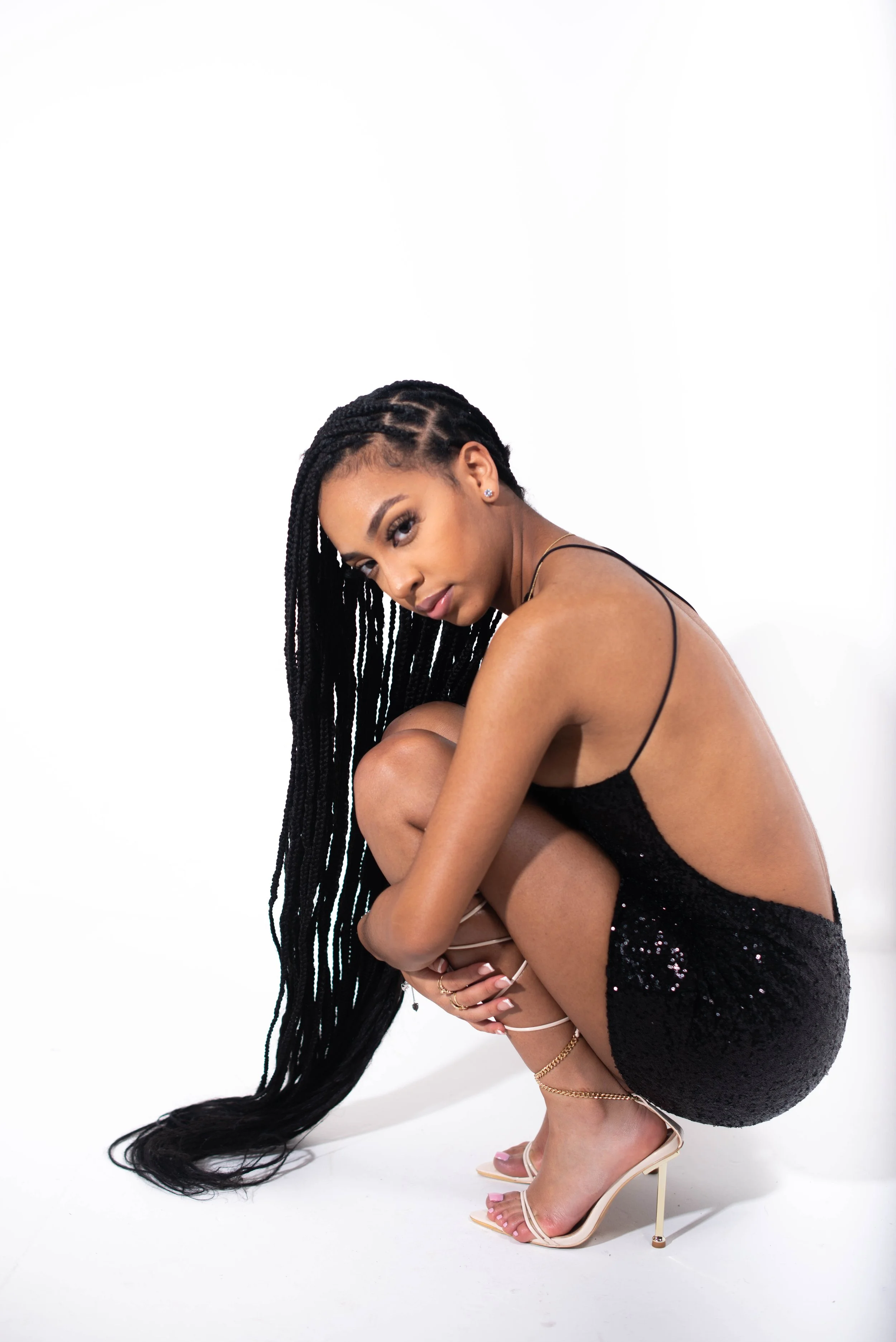 A woman with long black braids crouches down in a black sequined dress and nude high heels against a white background.