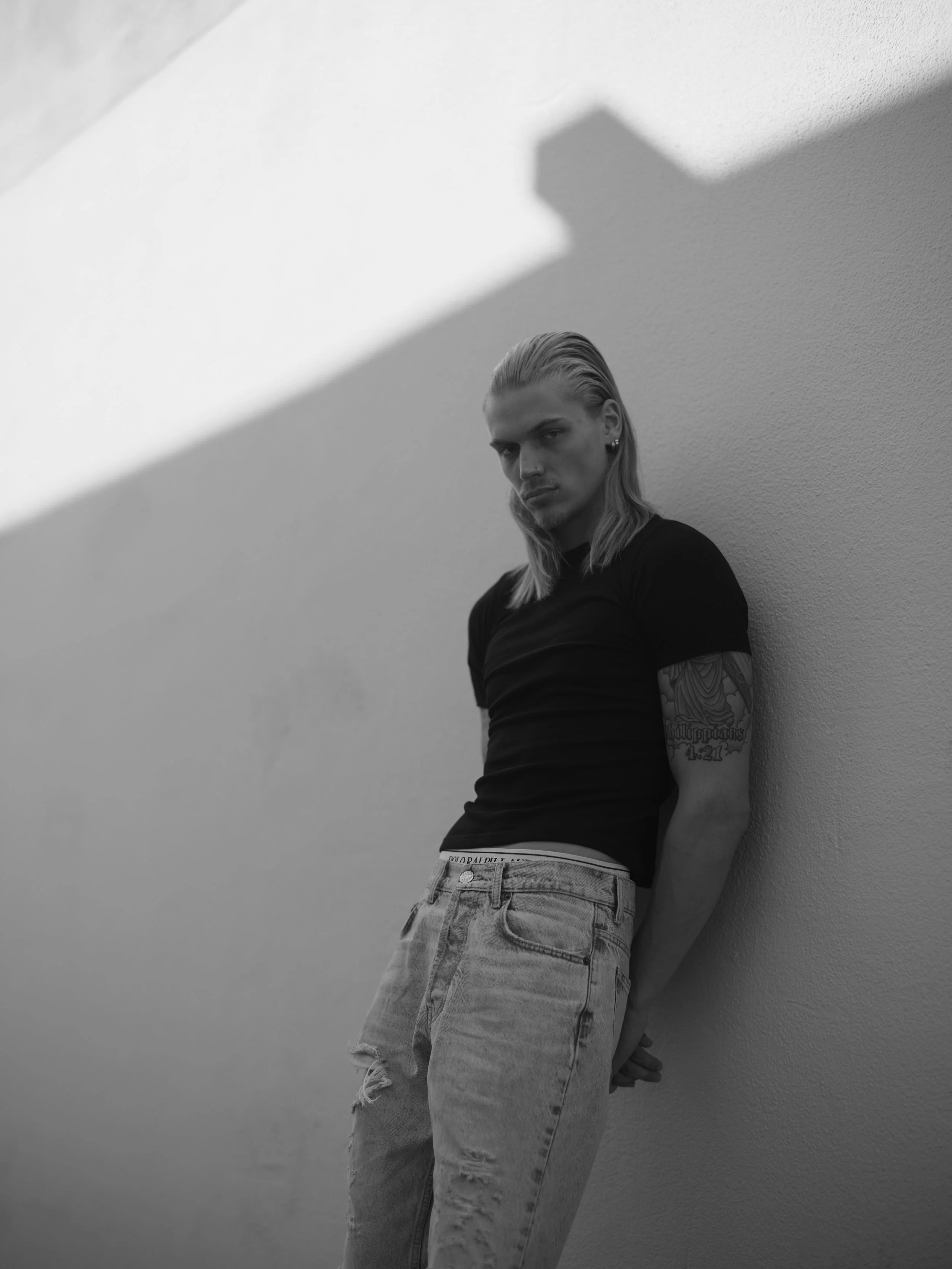 Black and white photo of a young man leaning against a wall, with a serious expression, long hair, tattoos on his arm, wearing a black t-shirt and ripped jeans.