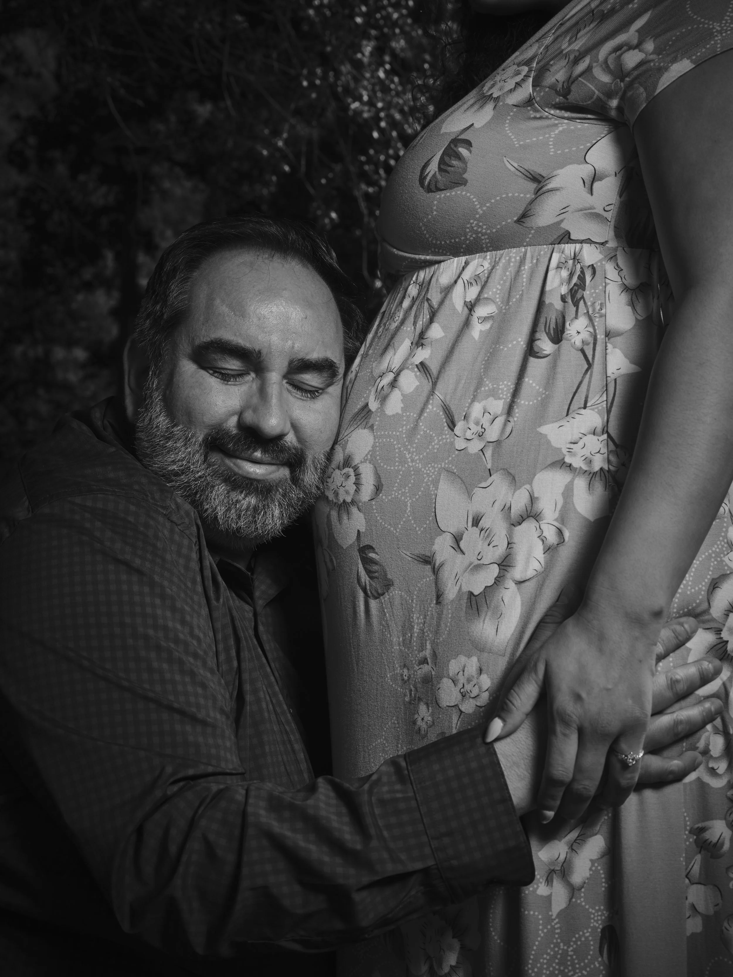 A man with a beard and closed eyes smiling, holding a pregnant woman's belly, which is wearing a floral dress. The photo is in black and white.