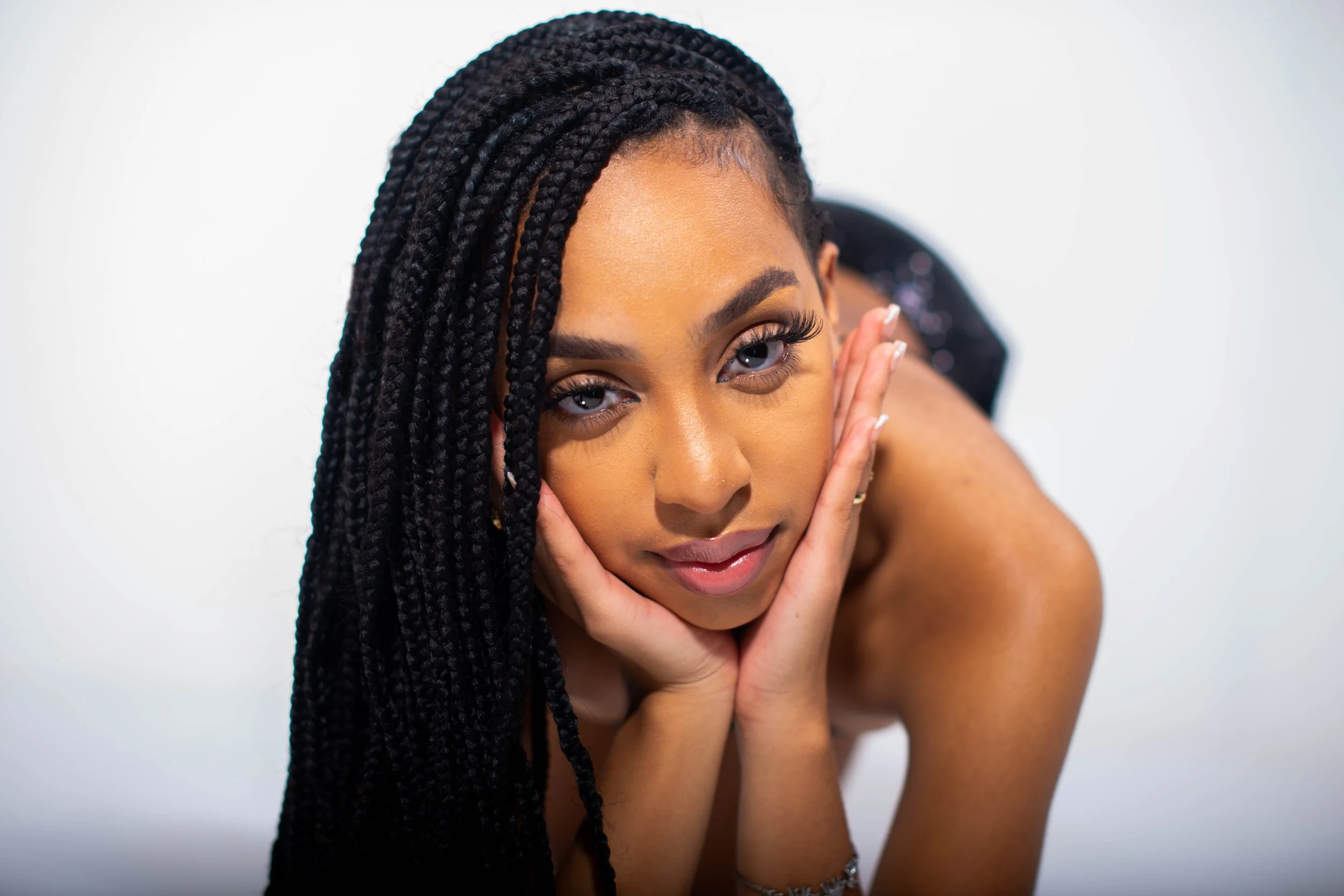 A woman with long, braided black hair resting her face in her hands, looking directly at the camera with a neutral expression, against a plain white background.
