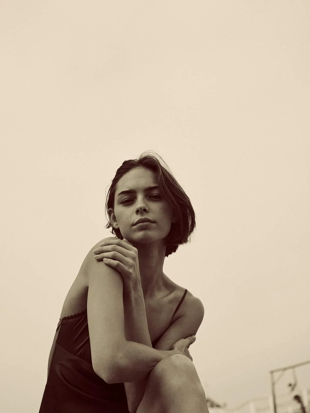 Black-and-white photo of a woman with shoulder-length hair, wearing a sleeveless top, looking into the camera with a confident expression, with her arms crossed over her knee, in an outdoor setting.