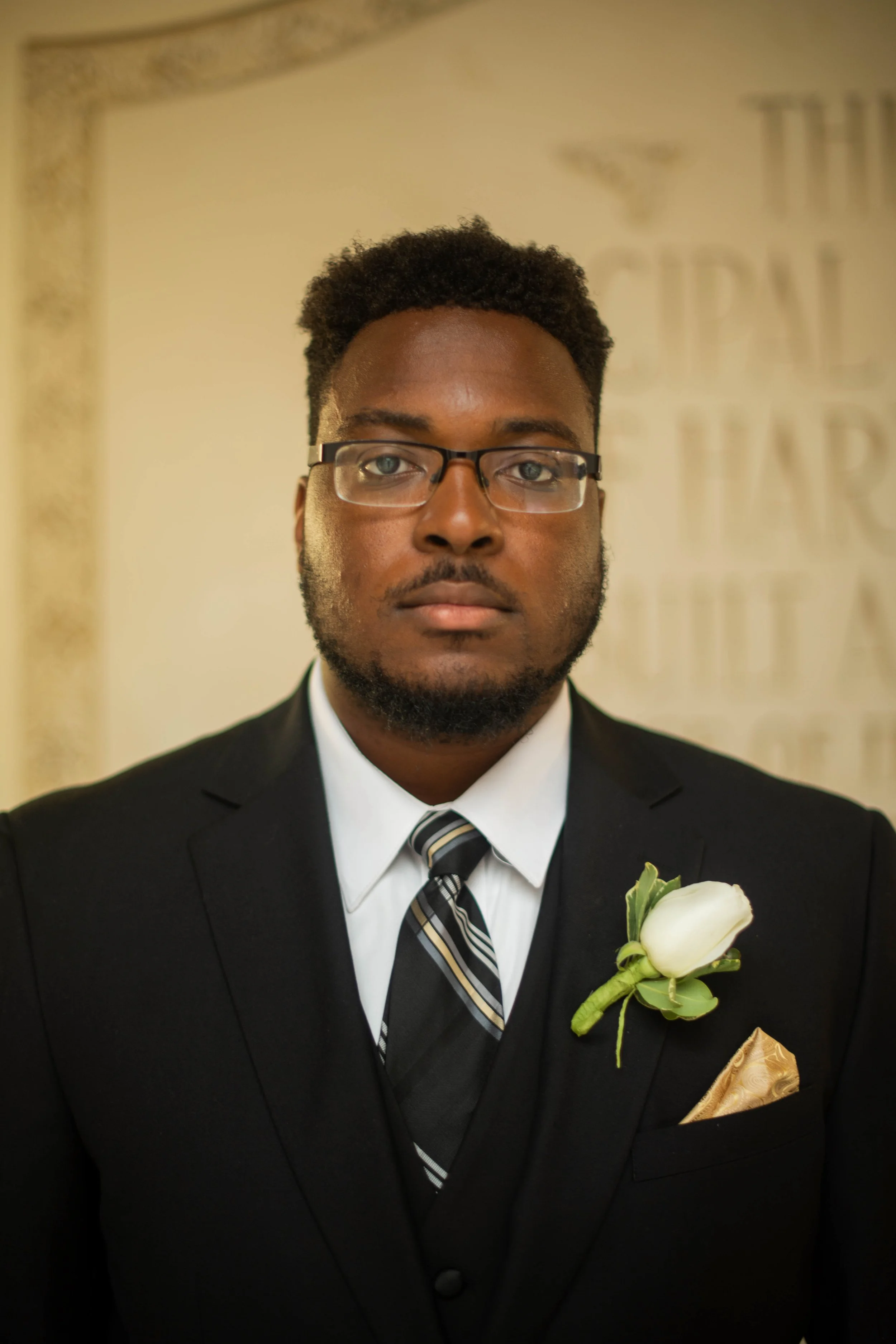 A man dressed in a black suit with a white shirt and striped tie, wearing glasses, and a white flower boutonniere on his lapel, standing indoors.