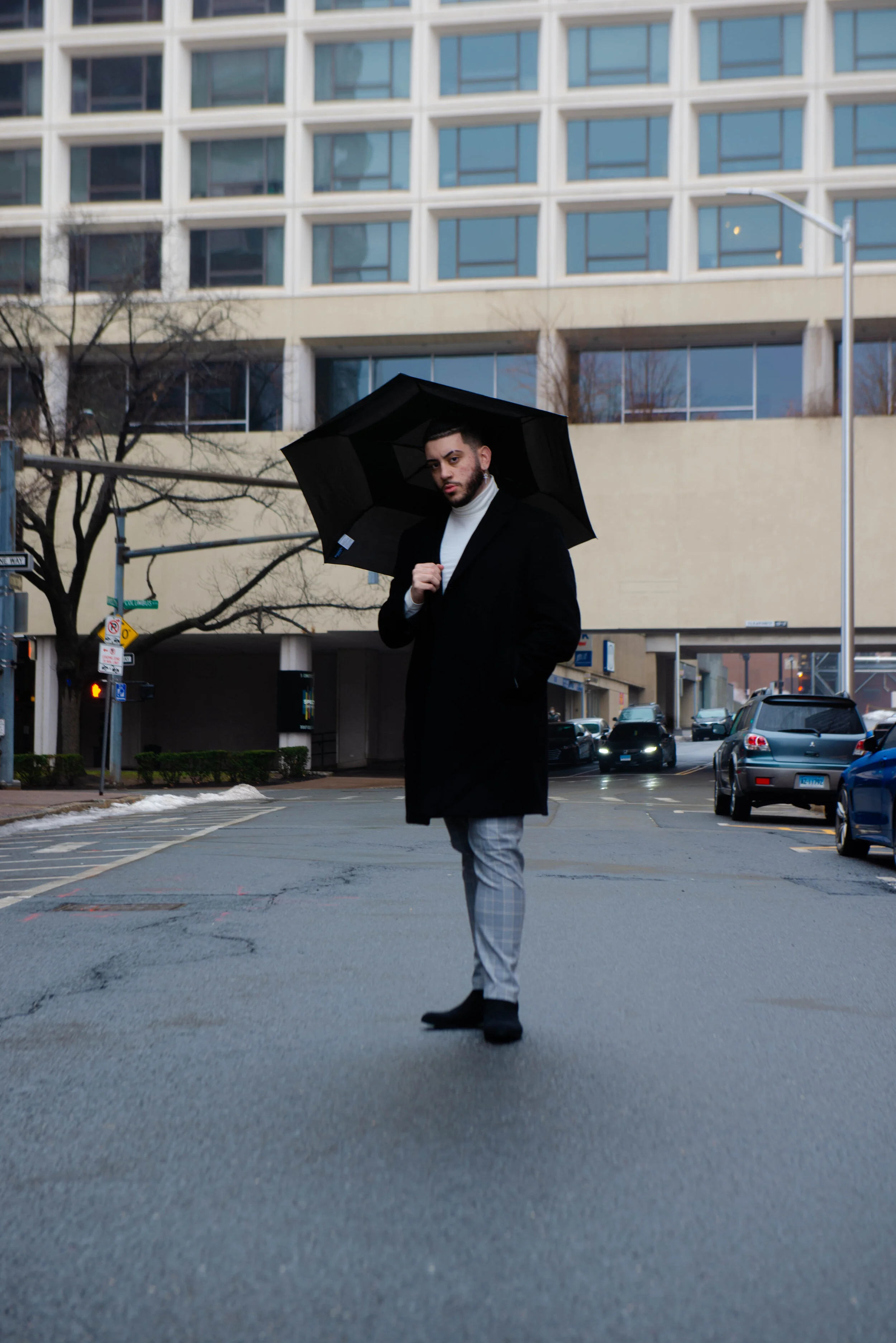 Young man standing on city street holding a black umbrella, wearing a black coat, white turtleneck, and plaid pants, with tall buildings and parked cars in the background.