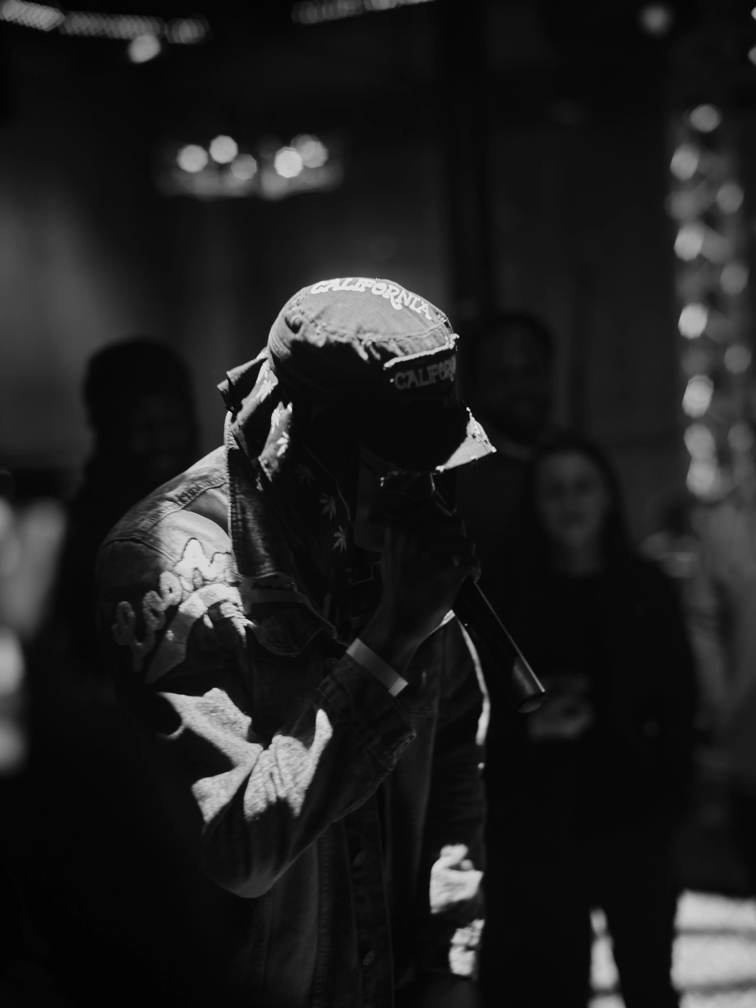 A person wearing a jacket and a cap labeled 'California' holding a microphone, with several people in the background. Black and white photo.