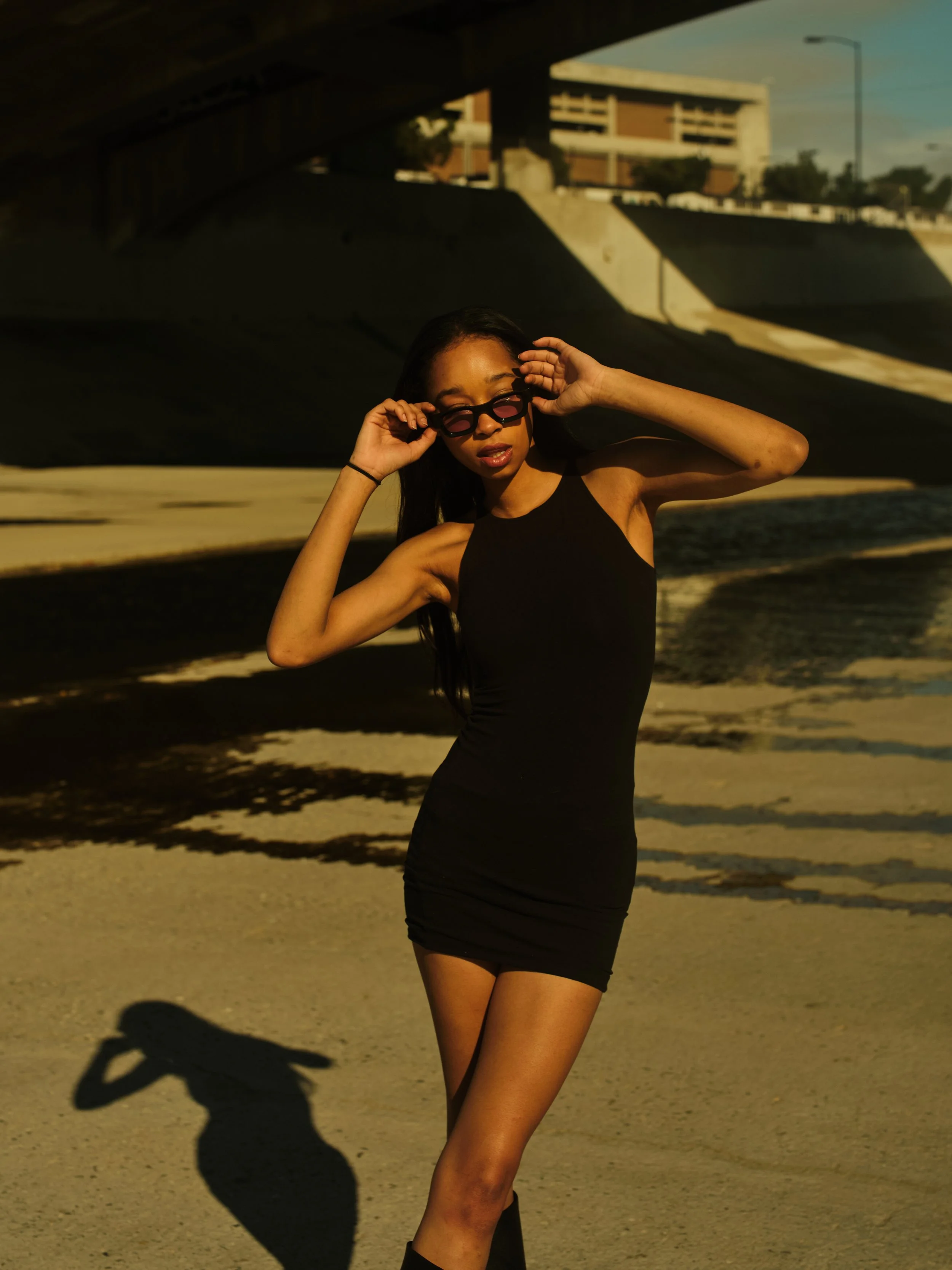 A woman in a form-fitting black dress and sunglasses striking a pose outdoors, with a large shadow cast on the ground and an urban bridge and buildings in the background.