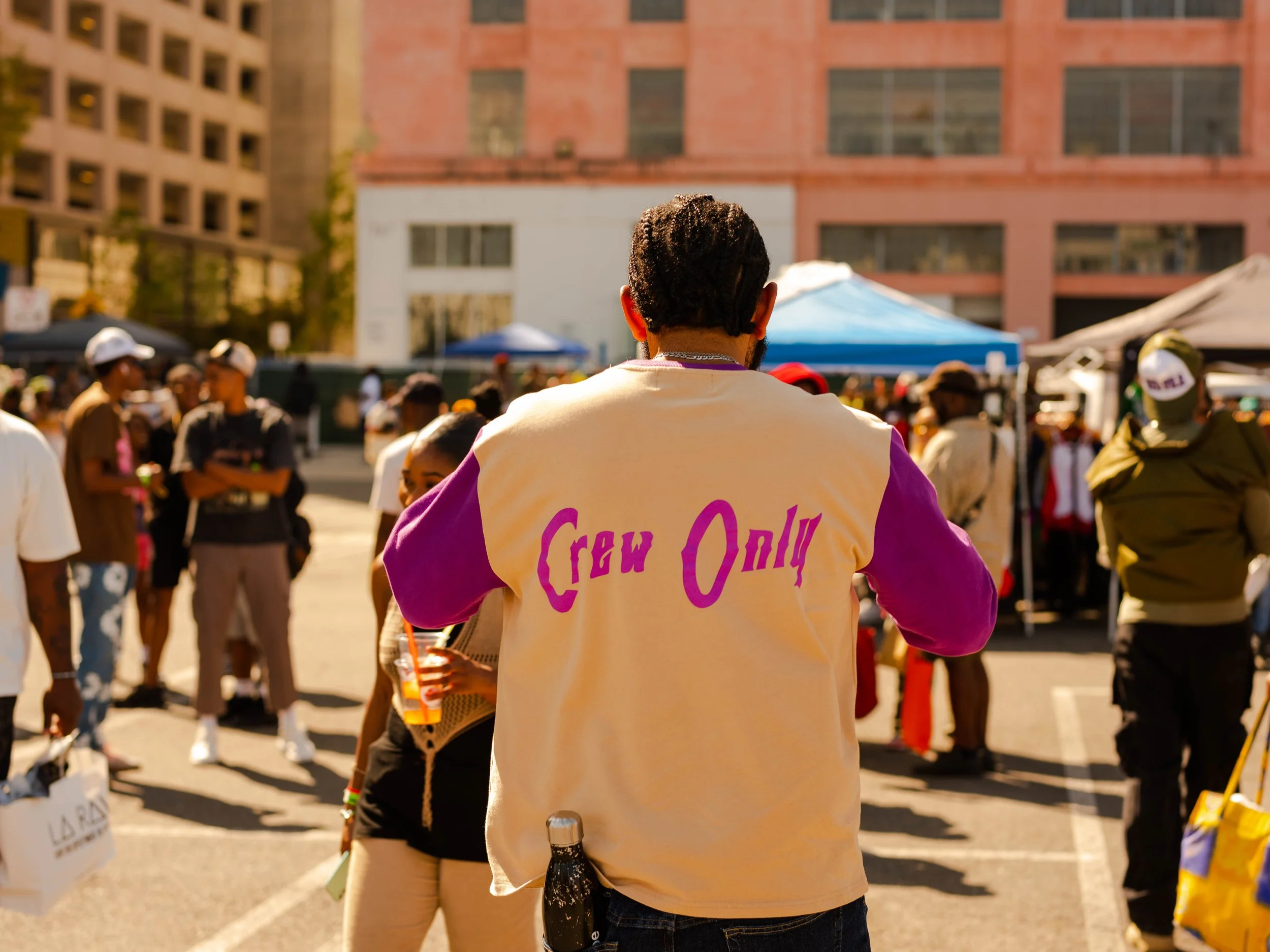 Person with a shirt that reads 'Crow Only' seen from the back in a crowded outdoor event with tents and many people