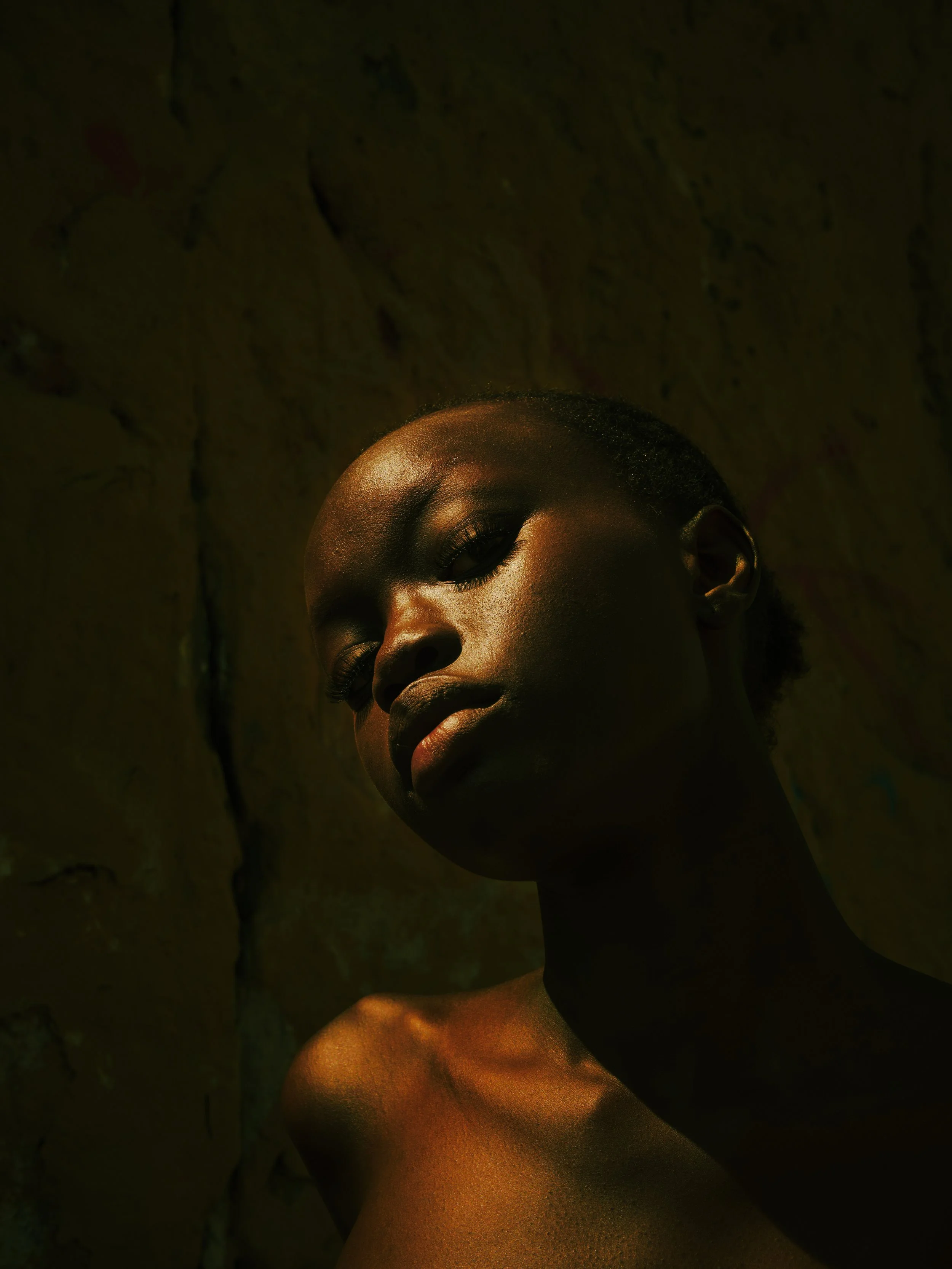 A portrait of a young woman with dark skin and short hair, illuminated by warm light, with a textured wall in the background.