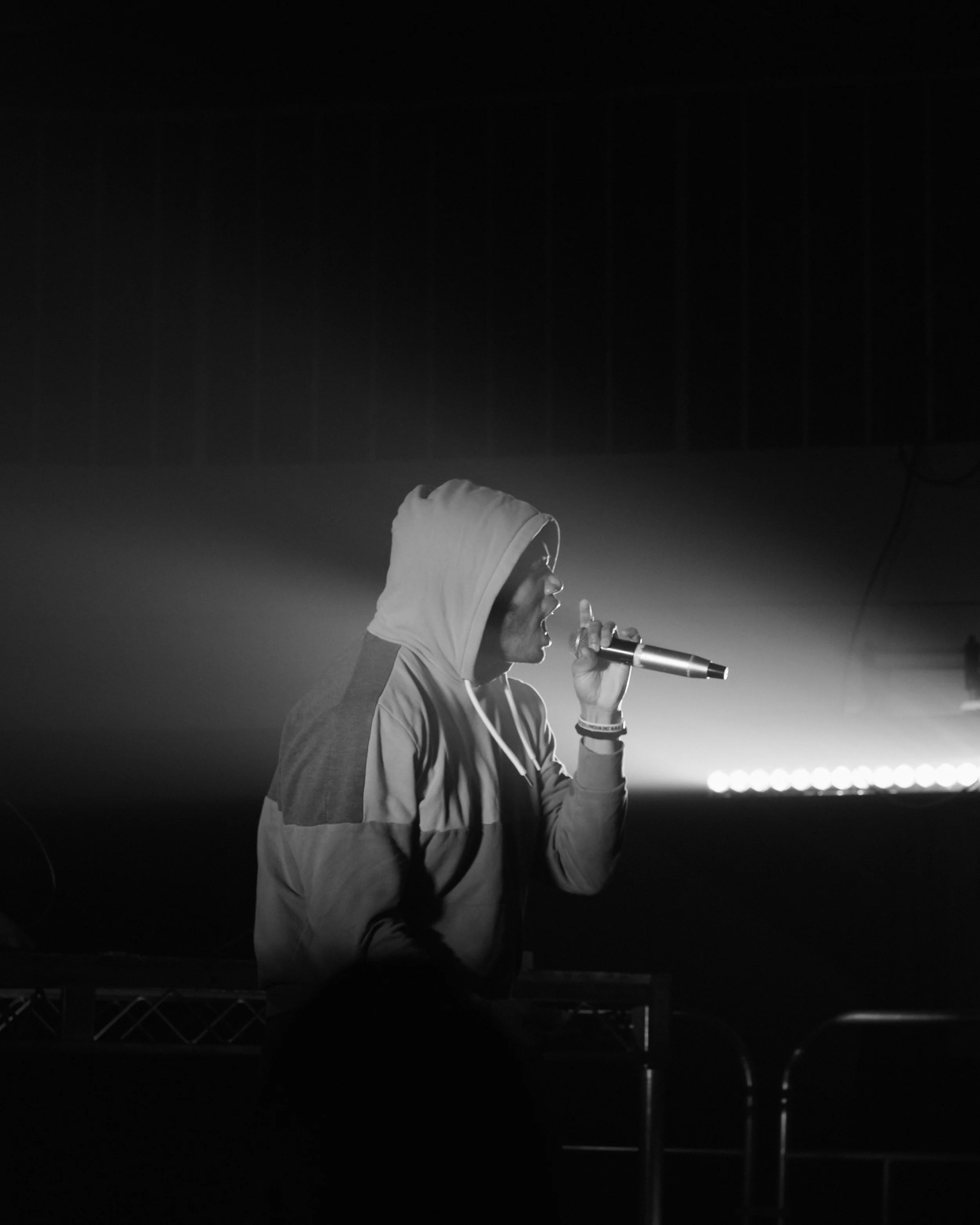 A person in a hoodie holding a microphone, performing on stage with a spotlight on them.