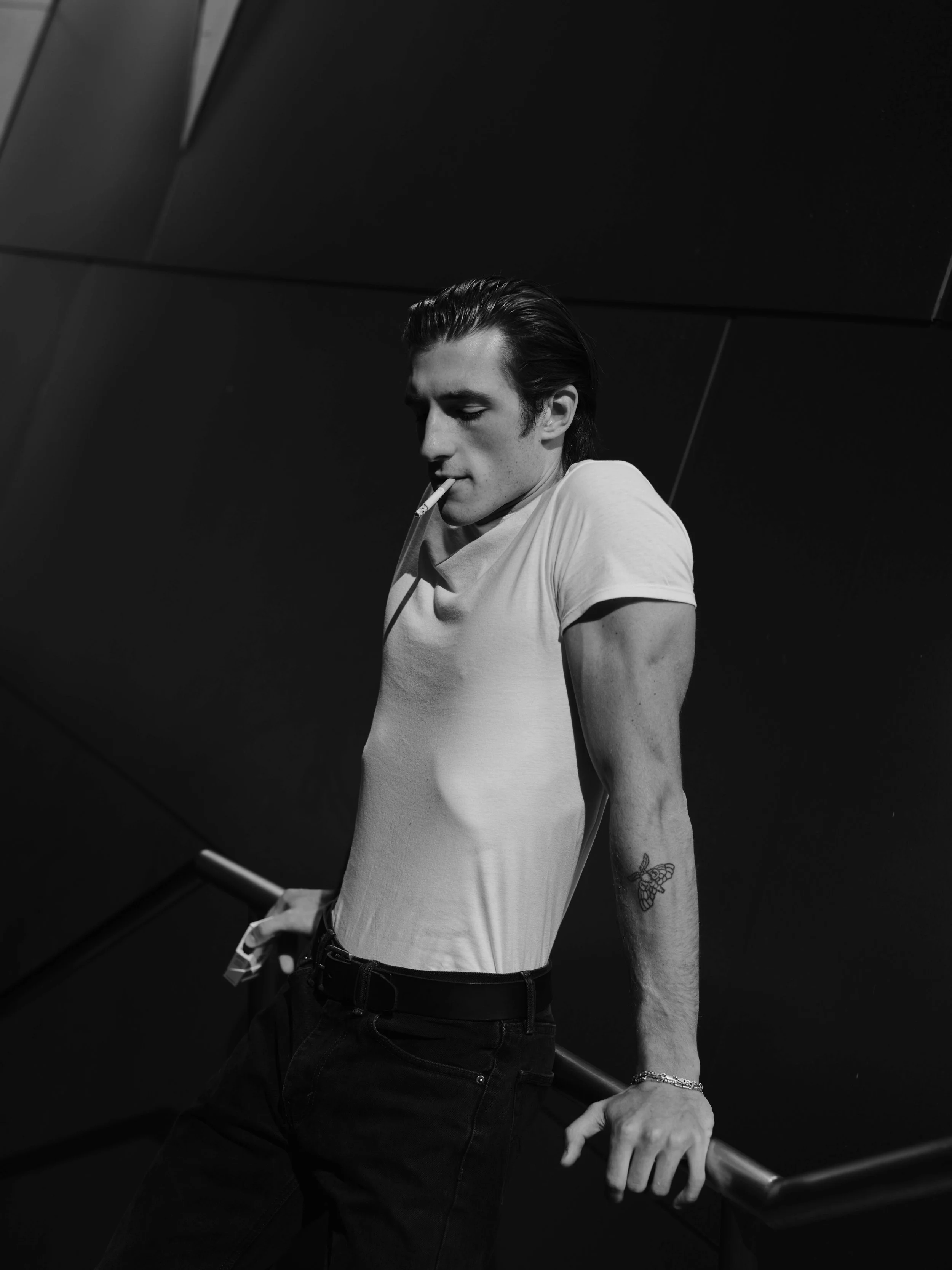 A black and white photo of a young man with slicked-back hair, wearing a tight white t-shirt and dark jeans, holding onto a railing with one hand and smoking a cigarette with the other, with a tattoo of a bee on his arm.