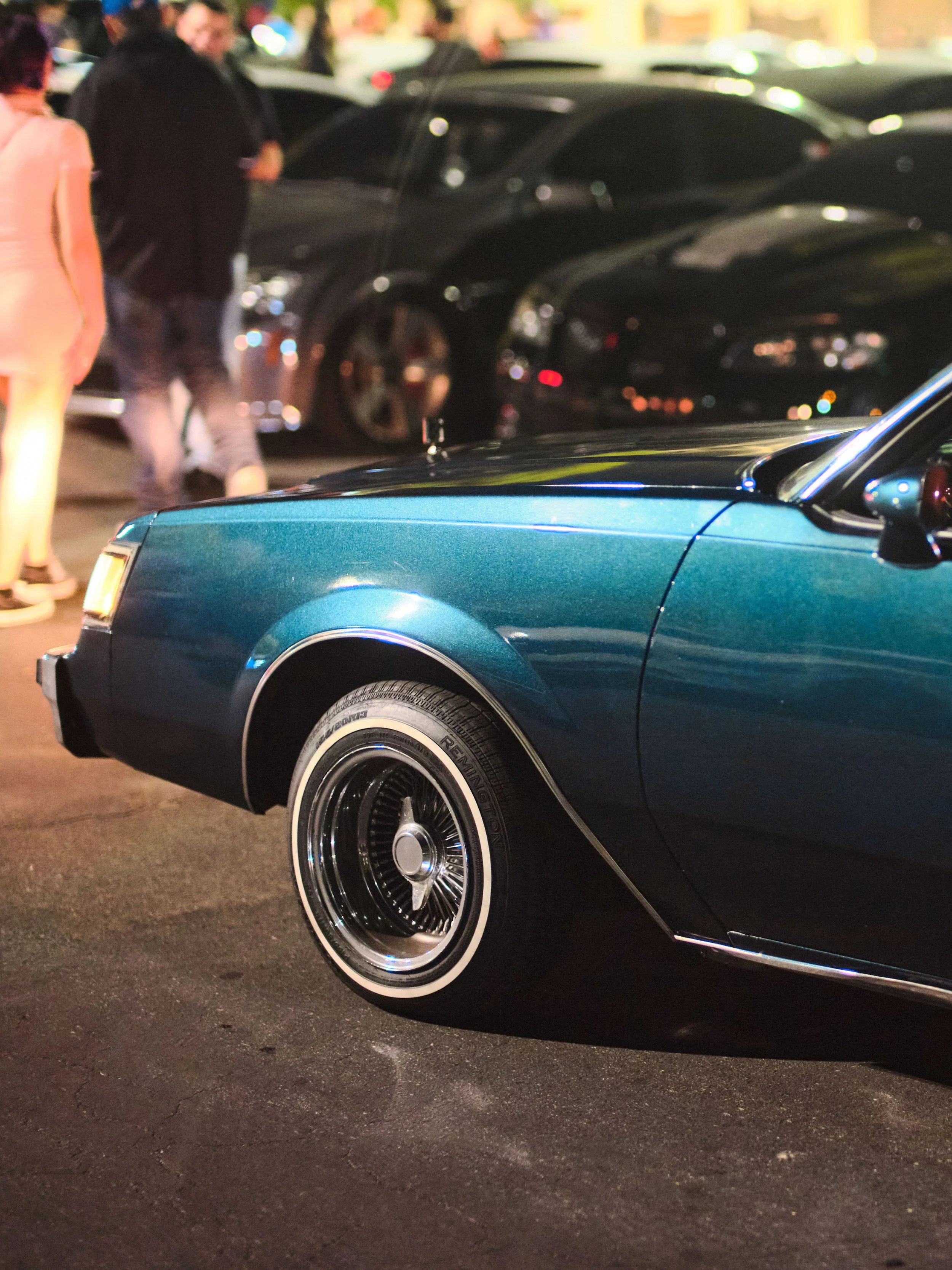 Close-up of a vintage blue car's front section with chrome detailing, parked in a lot at night with blurred people and other cars in the background.