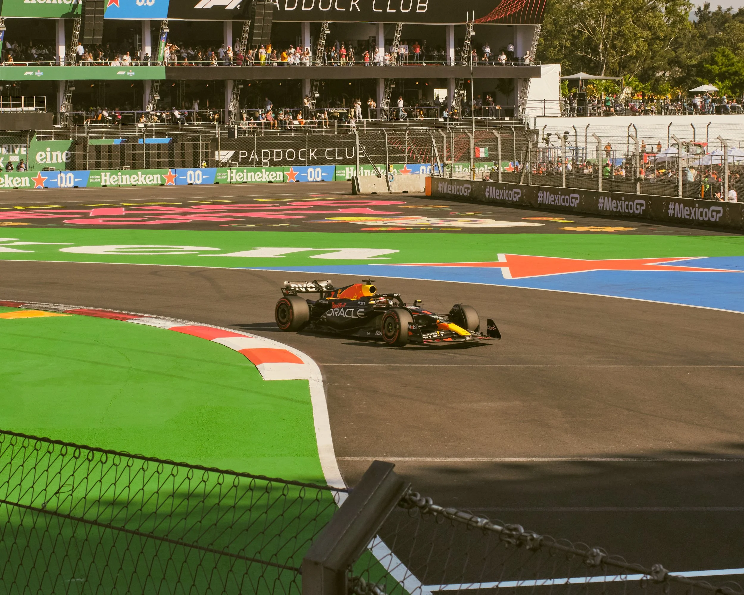A Formula 1 race car on a racetrack surrounded by colorful painted sections and barriers, with a grandstand filled with spectators in the background.
