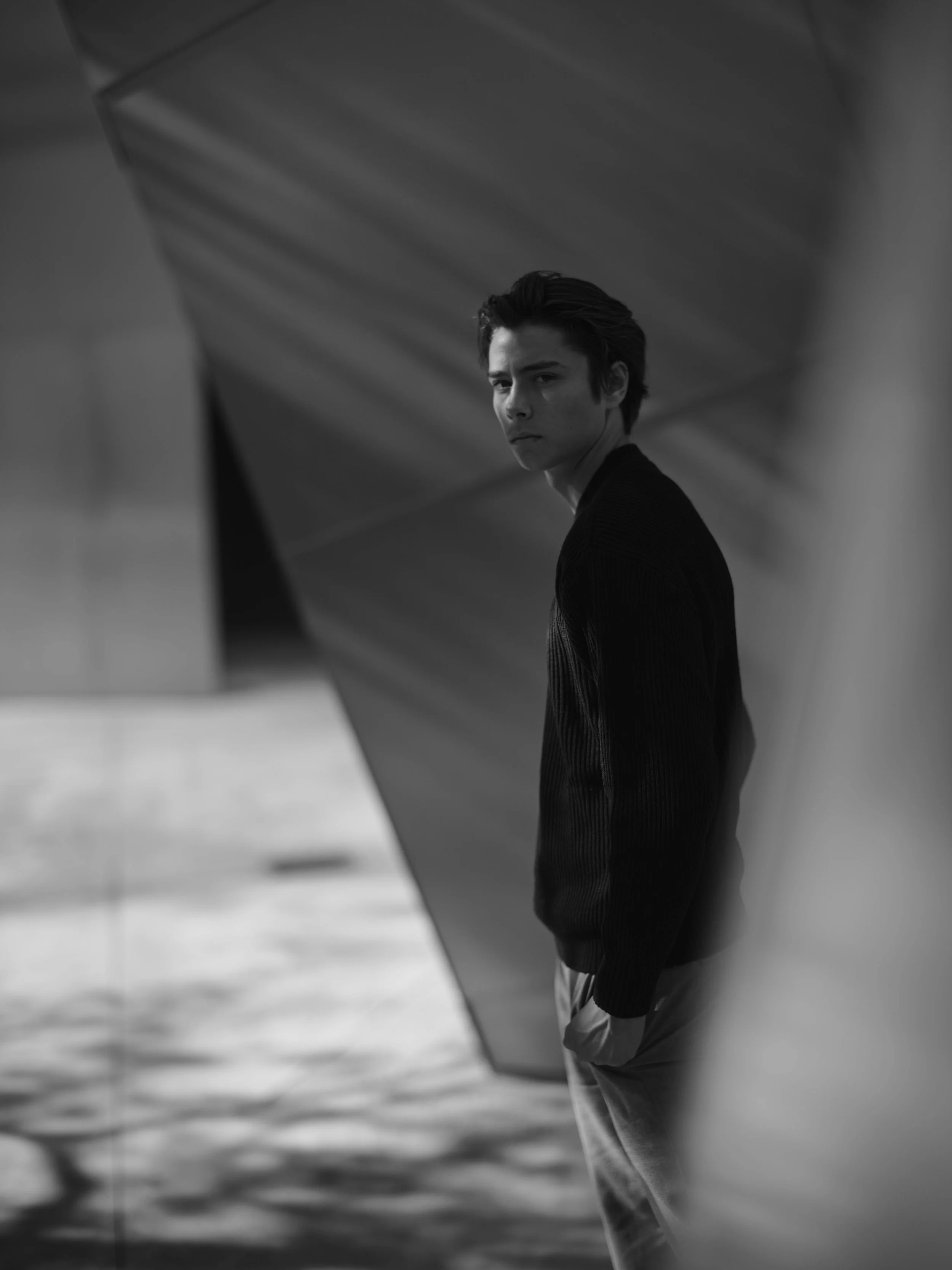 A young man with dark hair in a black sweater and light-colored pants stands against a modern architectural backdrop with geometric shapes, looking contemplatively at the camera in black and white.