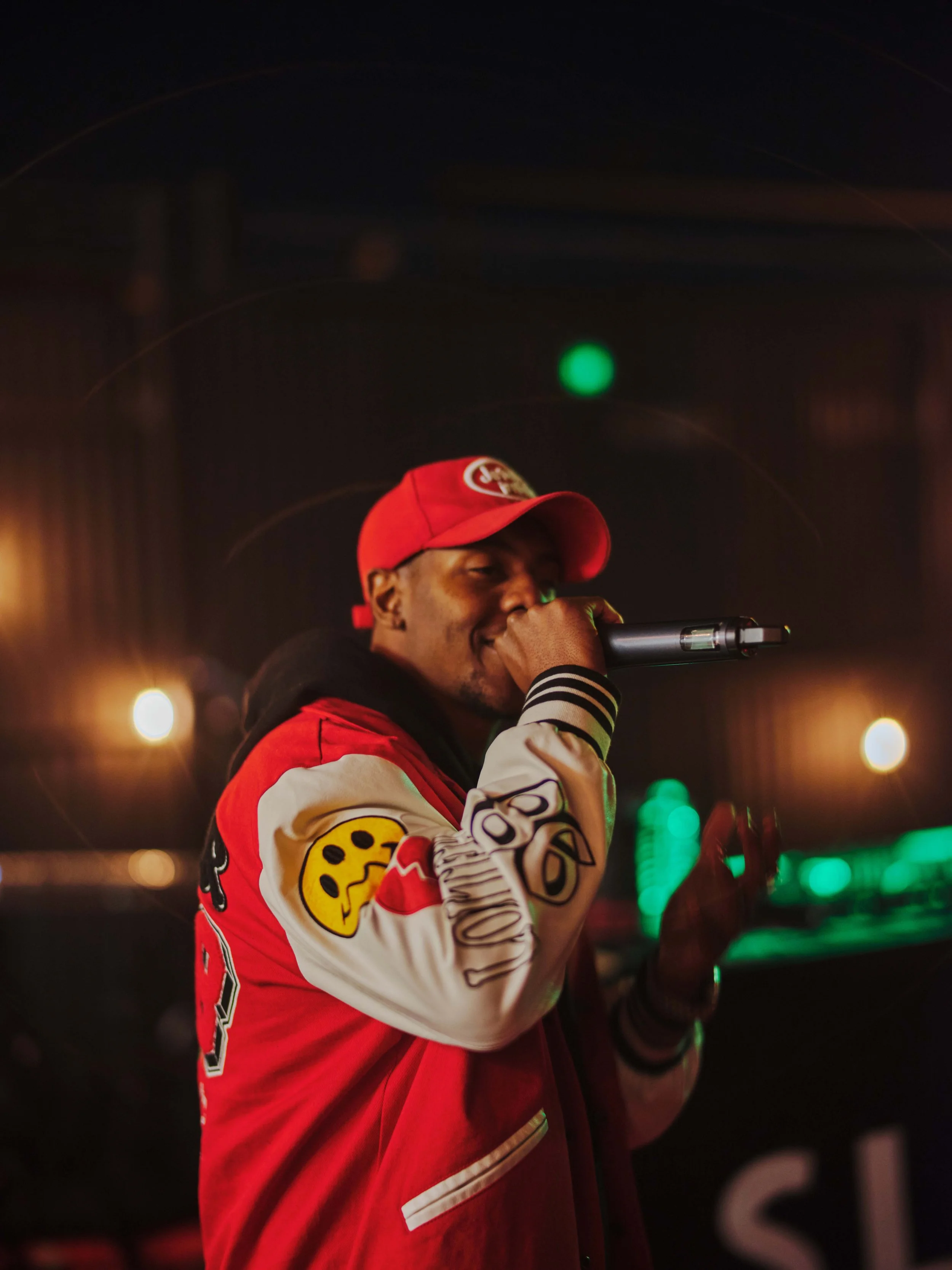 A man wearing a red cap and red jacket with embroidered patches is holding a microphone and performing on stage in a dimly lit setting.
