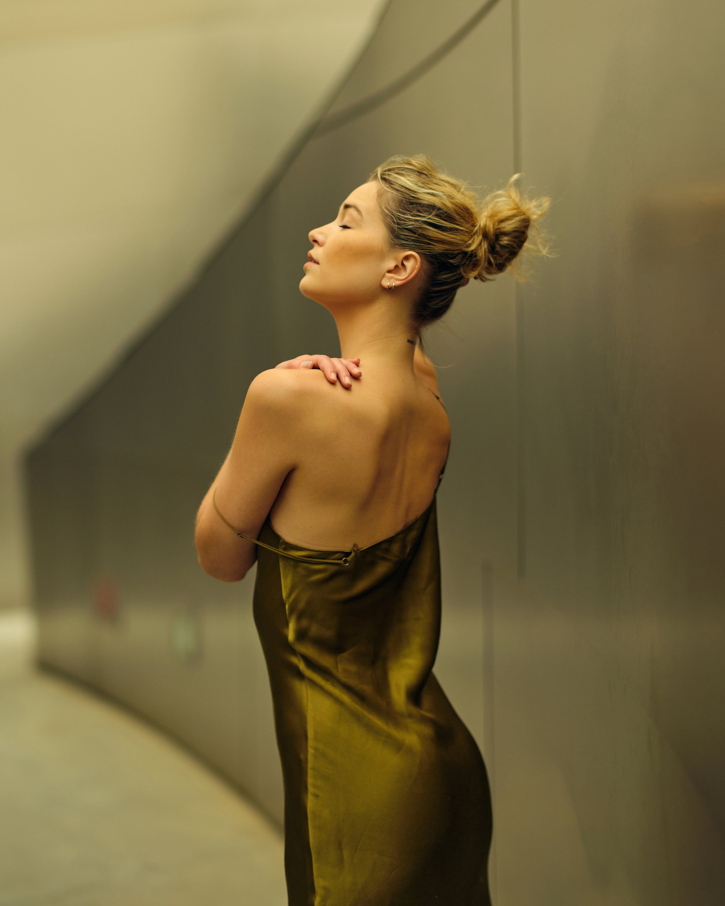 A woman with her eyes closed, standing with her arms crossed and holding her shoulders, wearing a satin dress gathered around her waist, with her hair in a messy bun, against a grey metallic wall.