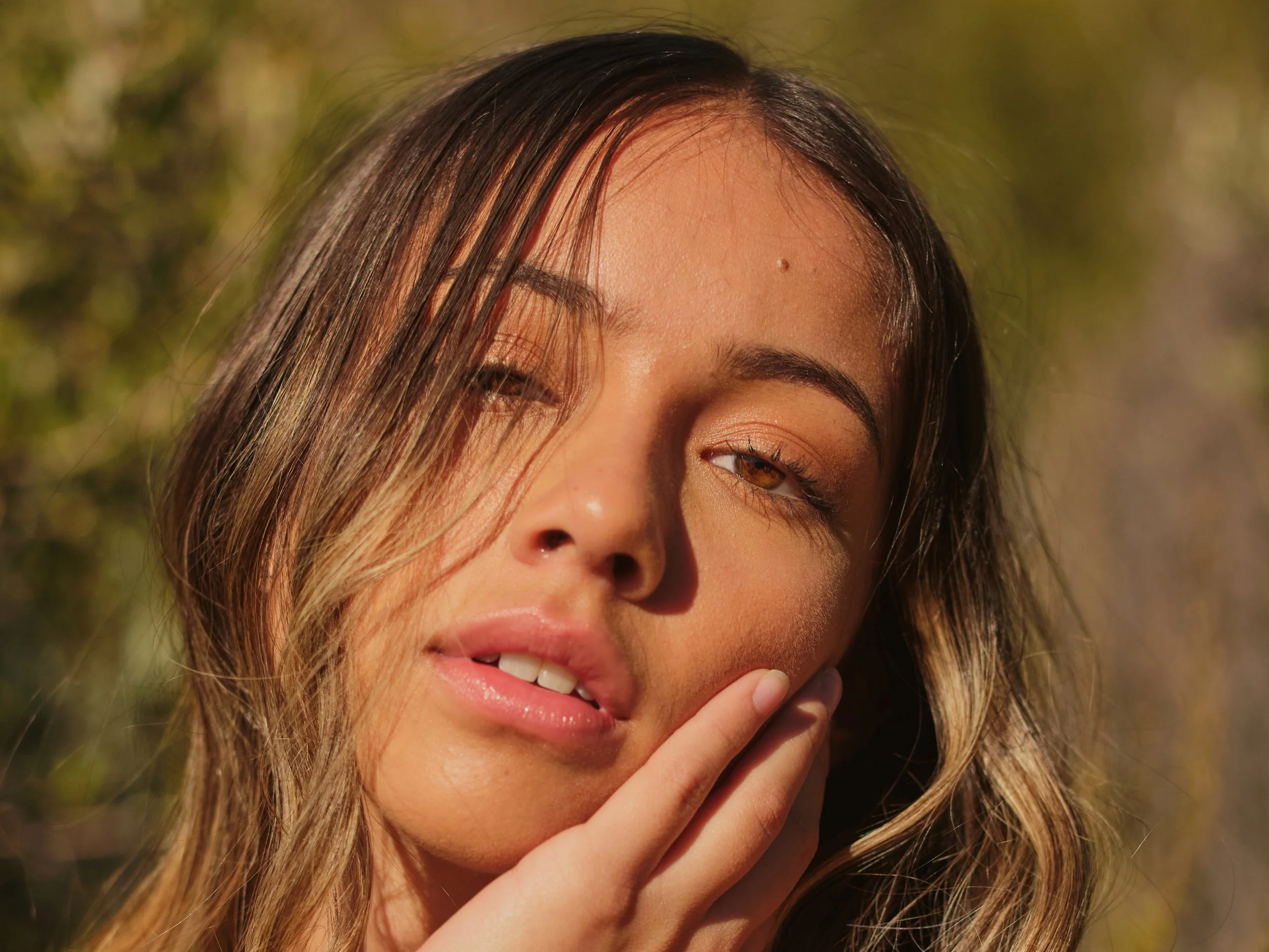 A young woman with long, wavy hair and light makeup posing outdoors in natural sunlight.
