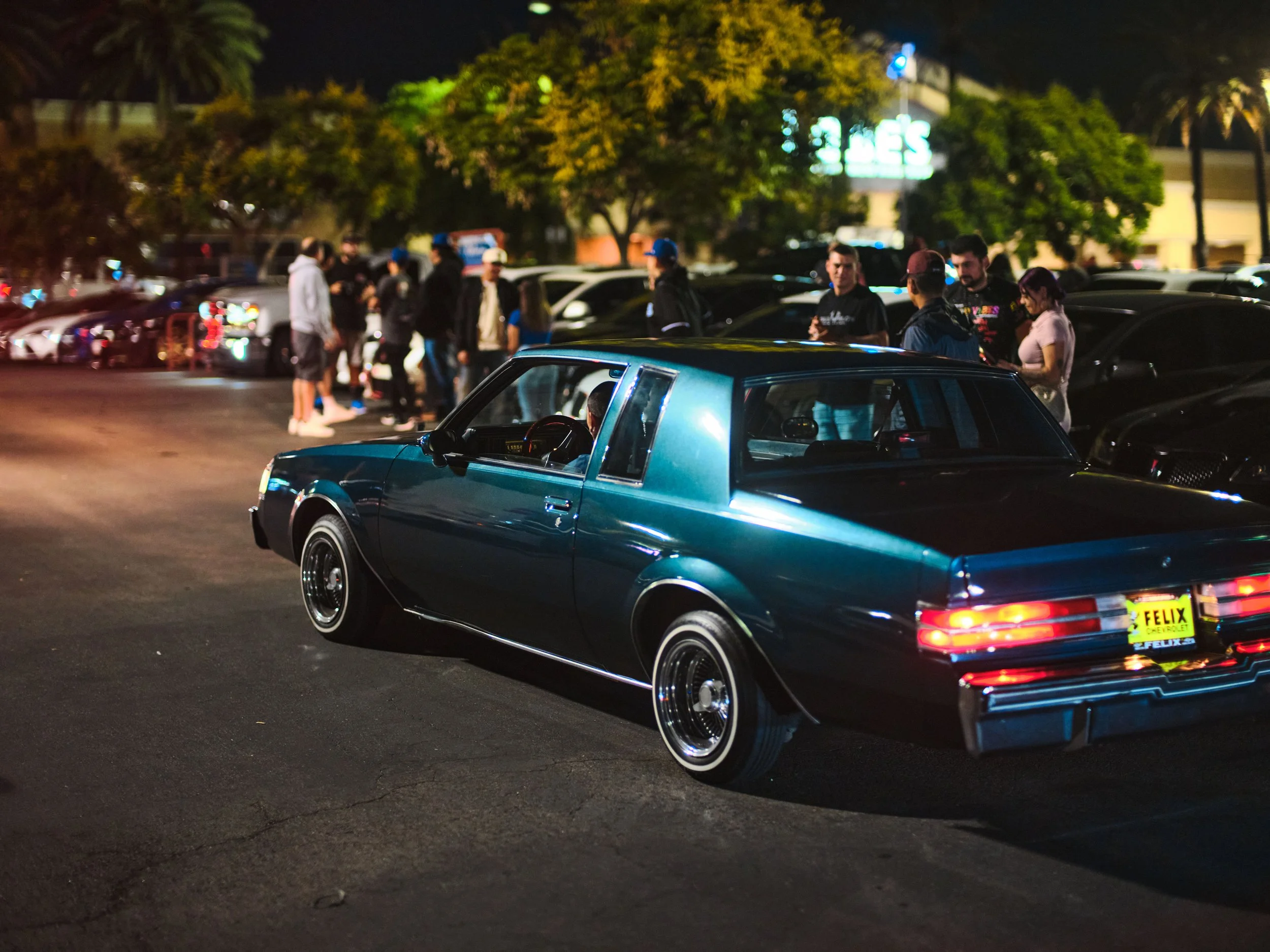 A vintage blue car parked in a lot at night with a group of people gathered in the background.